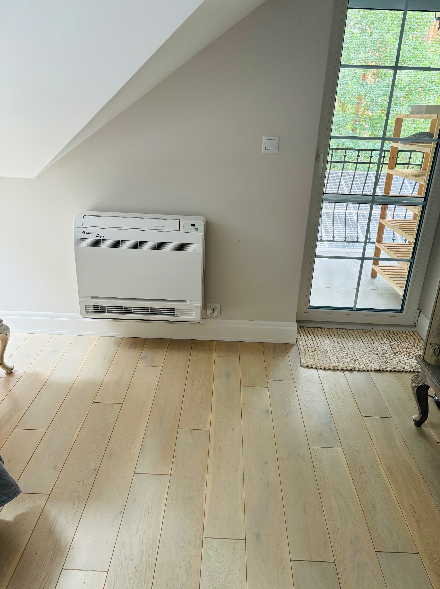 Biały klimatyzator podłogowy Gree Inverter w jasnym pomieszczeniu ze skośnym sufitem i oknem balkonowym. Widoczna podłoga z jasnych paneli.