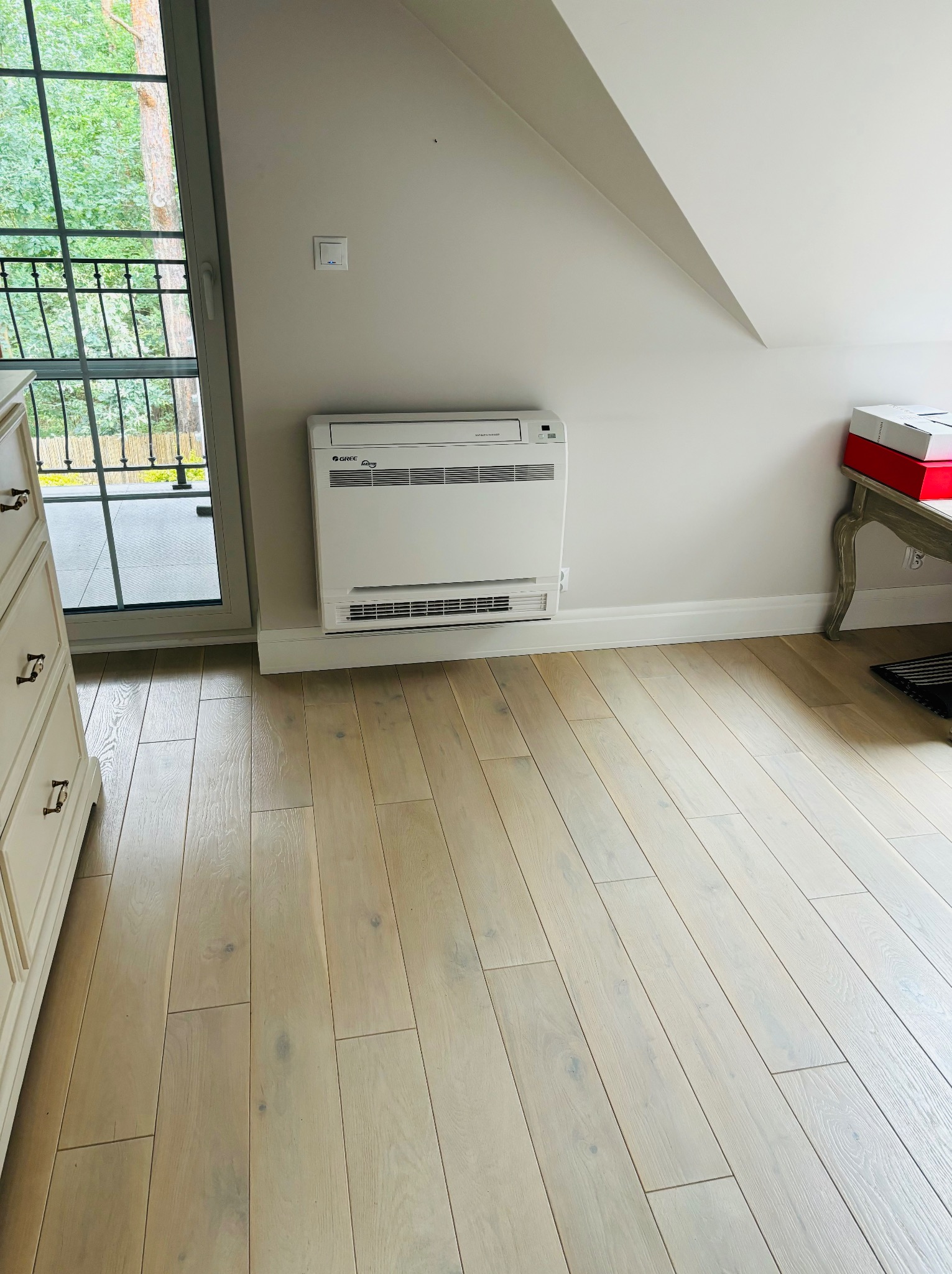 Biały, przypodłogowy klimatyzator Gree w jasnym pokoju na poddaszu z drewnianą podłogą, widok na balkon i drzewa za oknem.