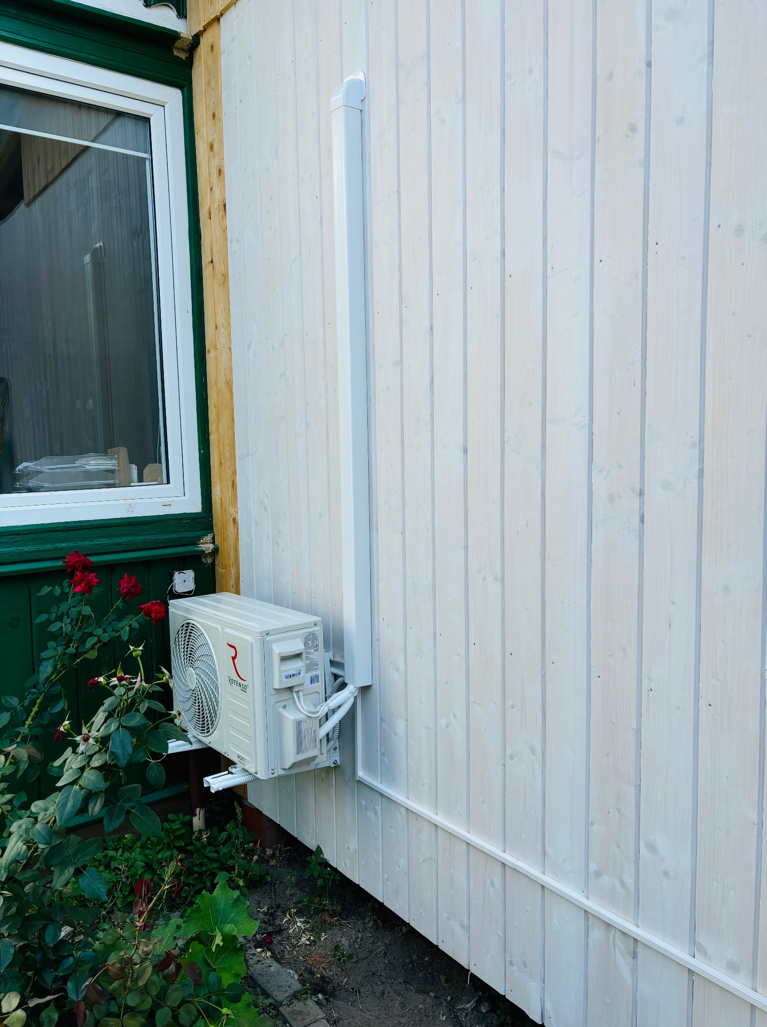 Zewnętrzna jednostka klimatyzacji Rotenso zamontowana na elewacji domu z białych desek, obok okna z zieloną ramą i krzewem róż.