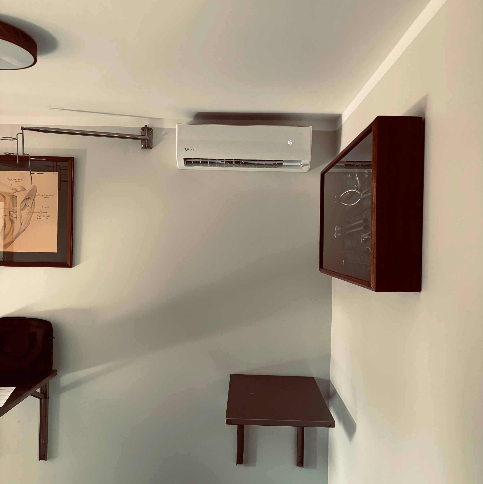 Biały klimatyzator ścienny Rotenso z wyświetlaczem temperatury 16 stopni Celsjusza, zamontowany w szarym pomieszczeniu z elementami dekoracyjnymi.