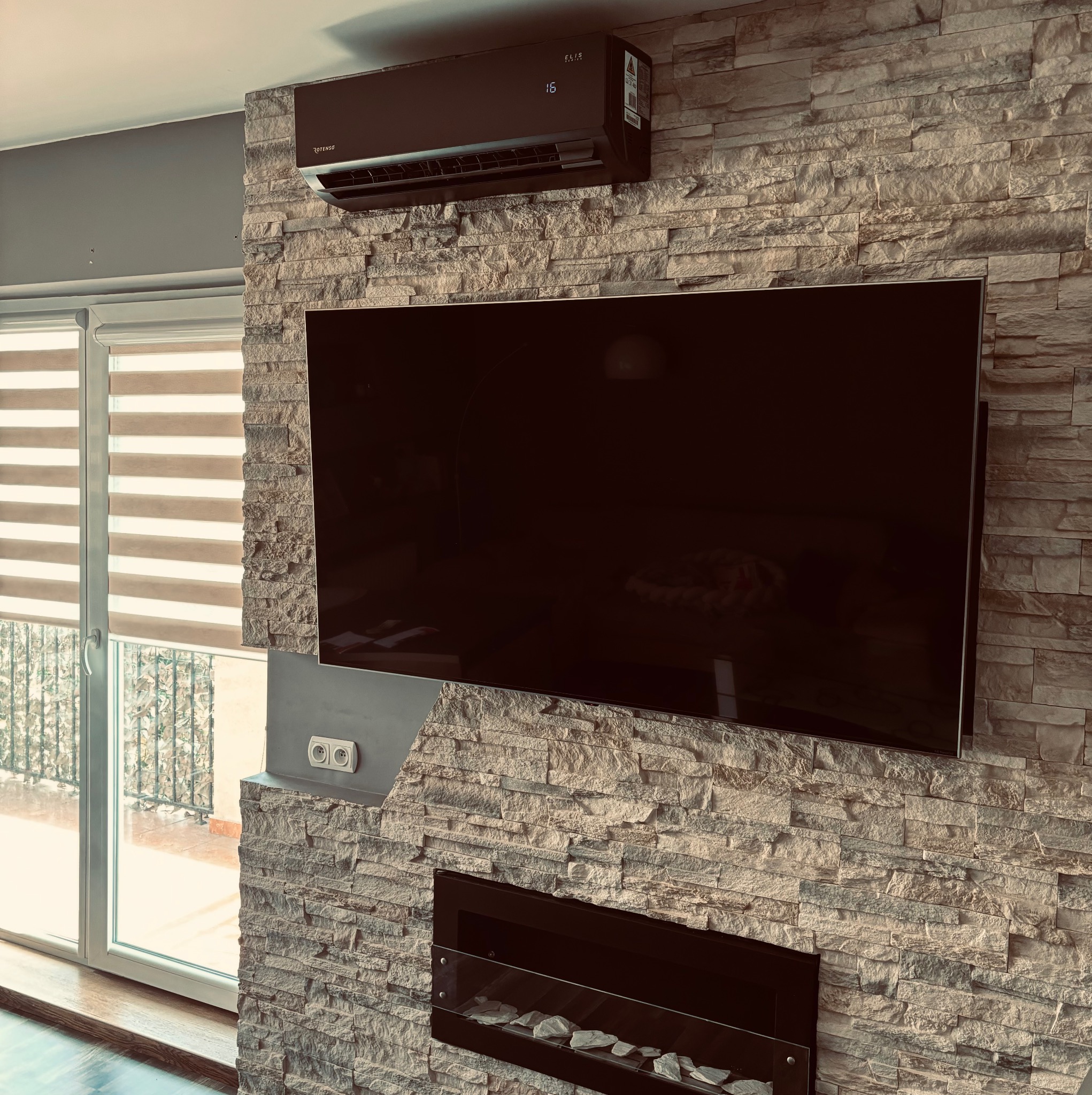 Nowoczesny klimatyzator Elis Series zamontowany w salonie w Warszawie, na ścianie z dekoracyjnej cegły, pod telewizorem i nad kominkiem.