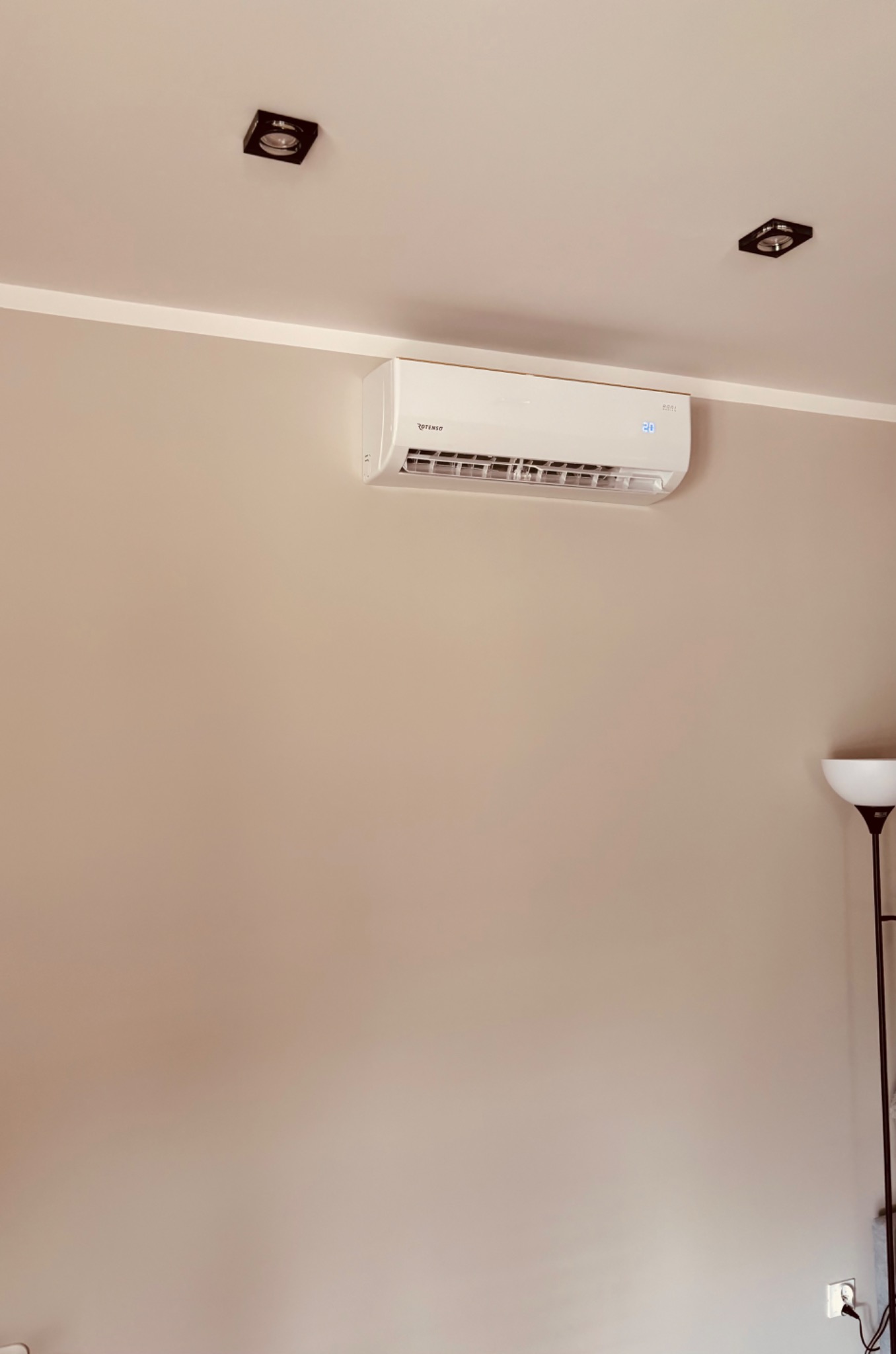 Biały klimatyzator Rotenso na ścianie w jasnym pokoju, widoczne dwa halogeny na suficie i lampa stojąca w rogu. Na wyświetlaczu klimatyzacji widoczna temperatura 20 stopni.