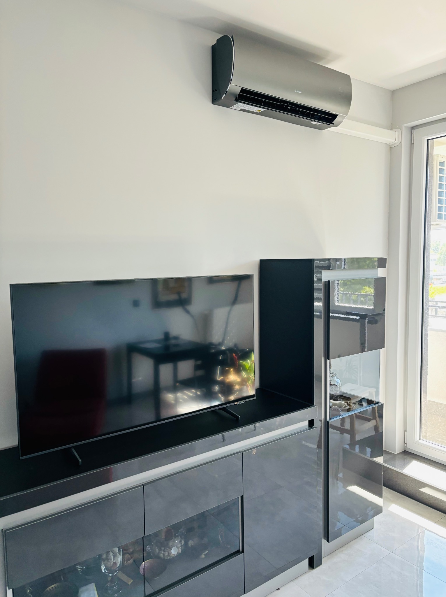 Szary klimatyzator ścienny Gree zamontowany w nowoczesnym salonie z błyszczącymi, grafitowymi meblami i dużym telewizorem. Widok na balkon.