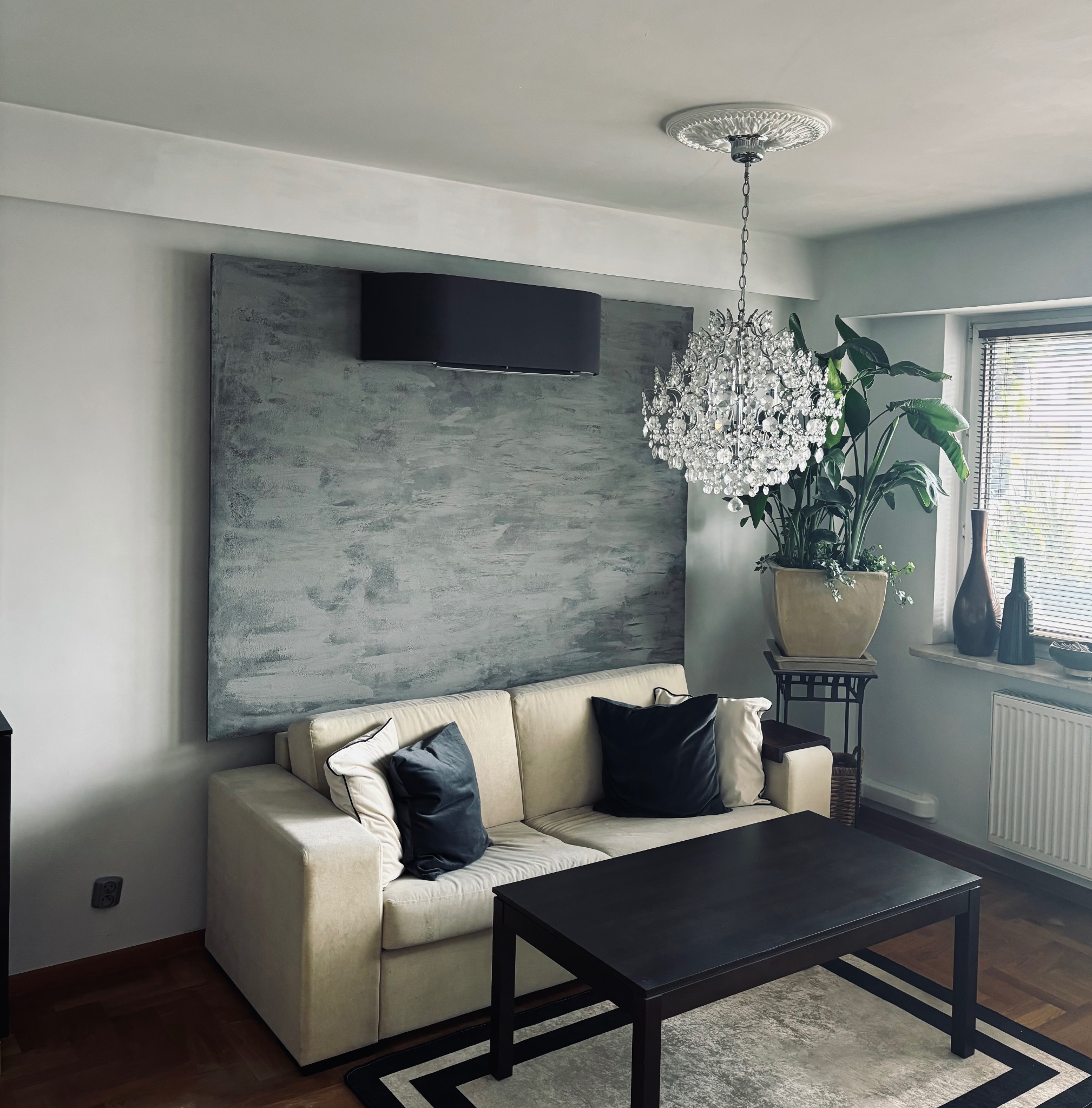 Elegancki salon z klimatyzatorem ściennym w kolorze czarnym nad sofą na tle szarej ściany. Wnętrze dopełnia kryształowy żyrandol i roślina doniczkowa.