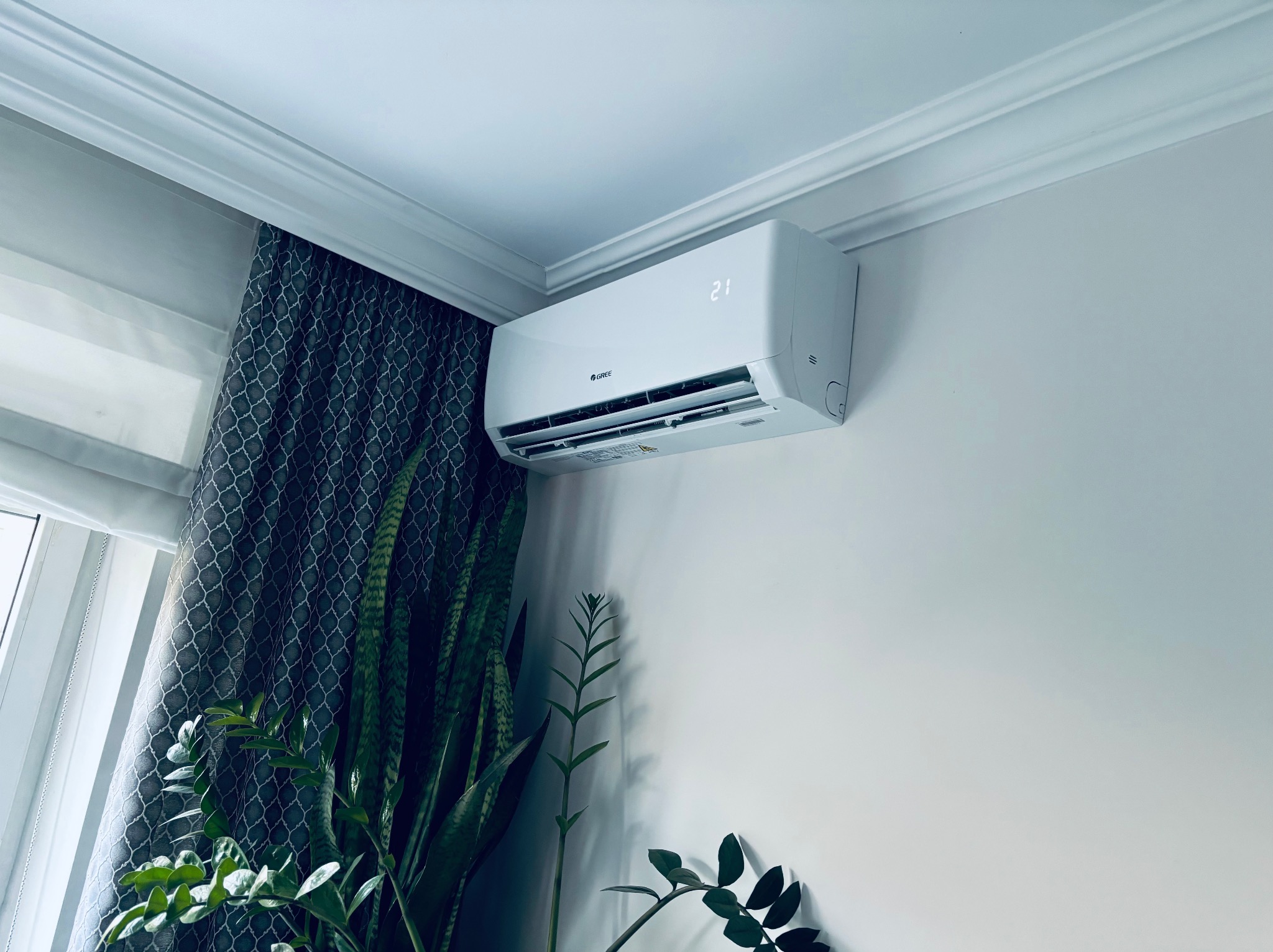 Biały klimatyzator marki Gree na ścianie w jasnym wnętrzu z roślinami i zasłoną. Wyświetlacz klimatyzatora pokazuje temperaturę 21 stopni.
