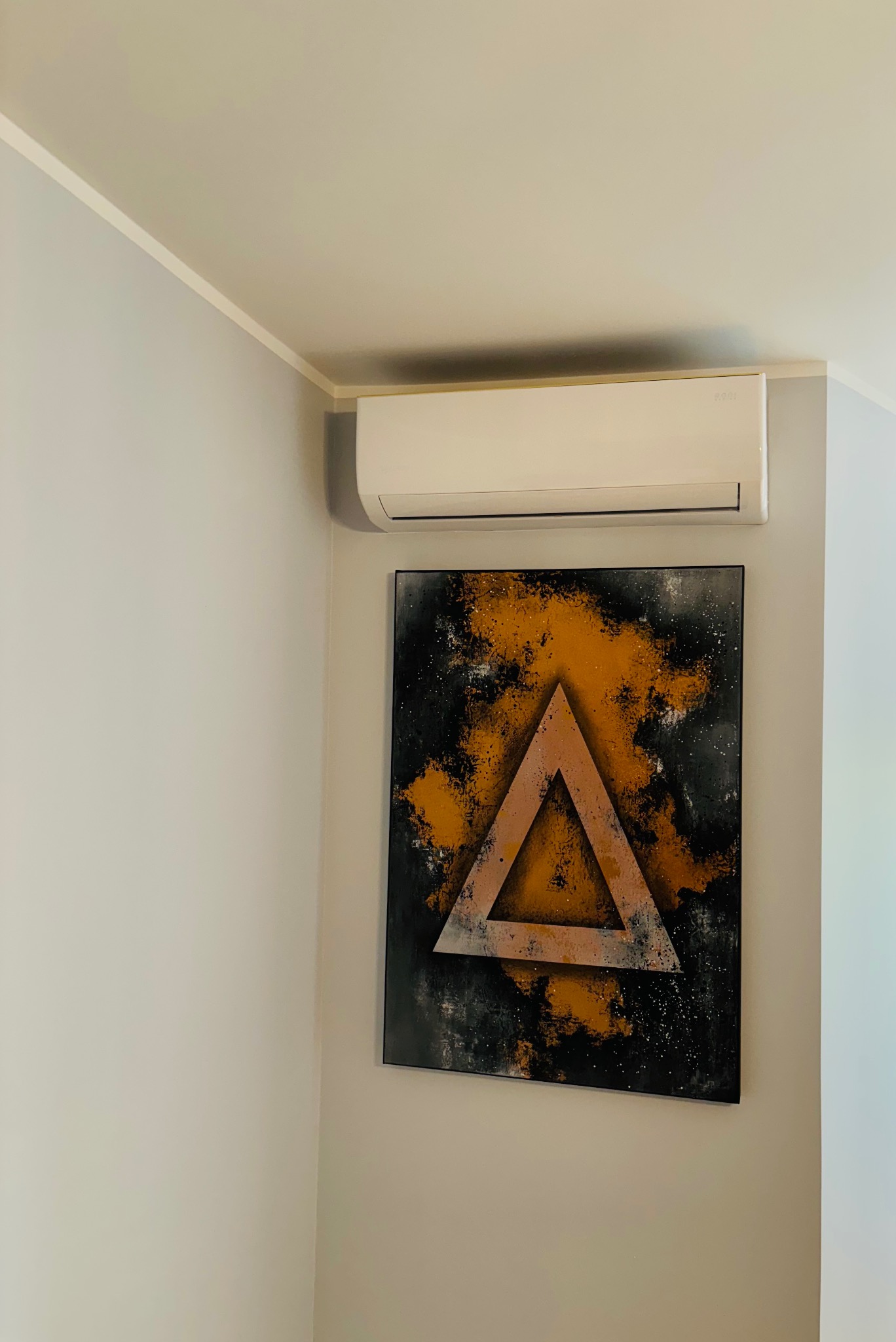 Klimatyzator ścienny zamontowany nad obrazem z motywem trójkąta w salonie o jasnych ścianach. Widok z dołu, podkreślający estetyczne dopasowanie urządzenia.