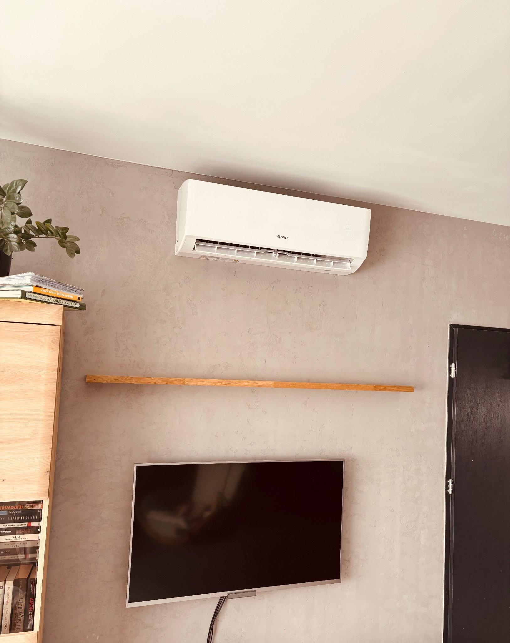 Biały klimatyzator Gree na ścianie o fakturze betonu, z półką i telewizorem. Widoczny fragment regału z książkami i kwiat doniczkowy. Jasne, naturalne światło.