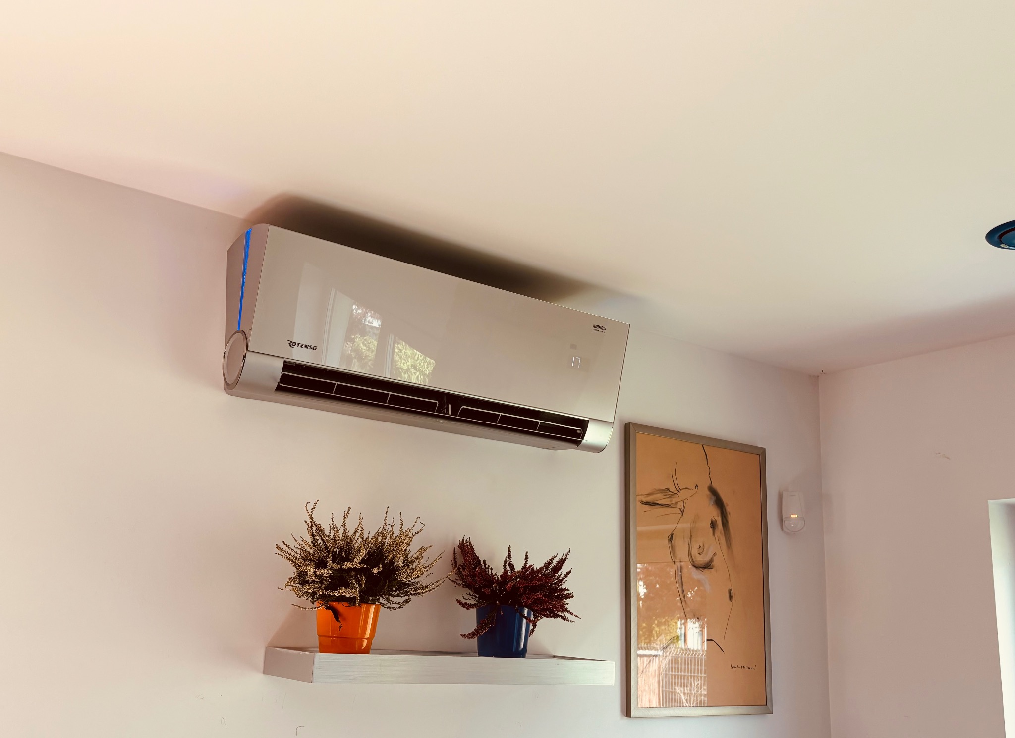 Elegancki klimatyzator Rotenso na białej ścianie, nad półką z kwiatami w kolorowych doniczkach, obok obraz w ramie. Nowoczesne wnętrze z klimatyzacją.