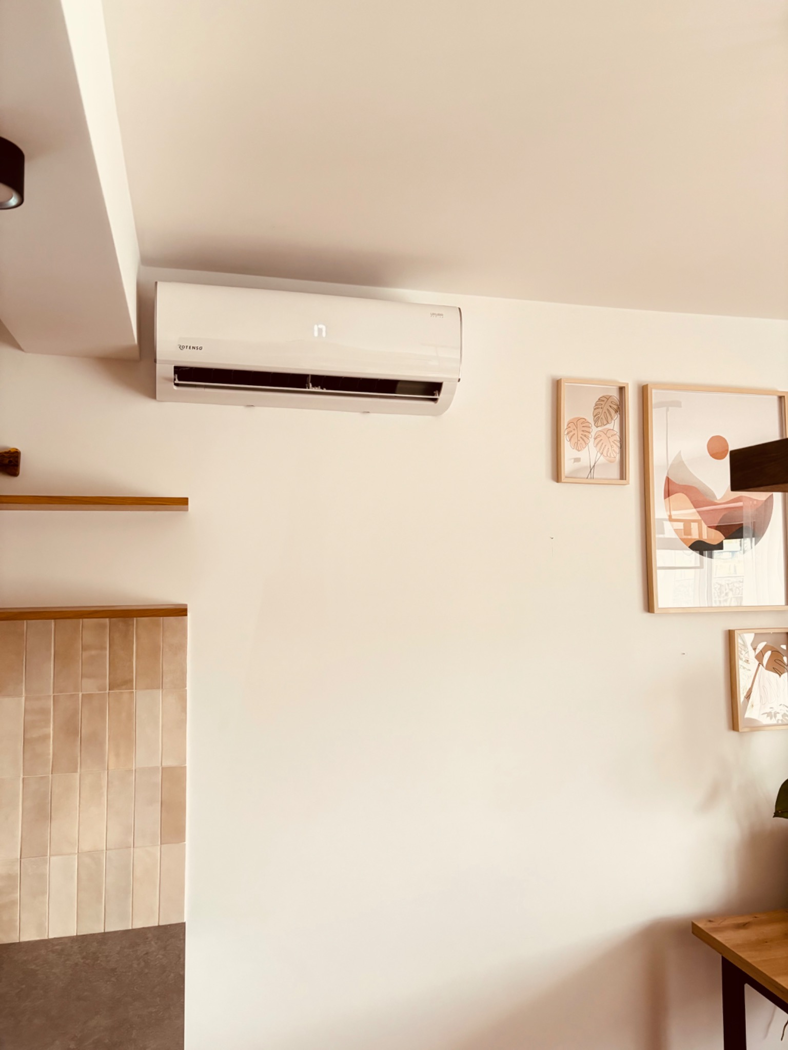 Klimatyzator Rotenso na ścianie w jasnym wnętrzu. Minimalistyczne obrazy w ramkach i drewniane półki. Widoczny fragment blatu i dekoracyjne płytki.