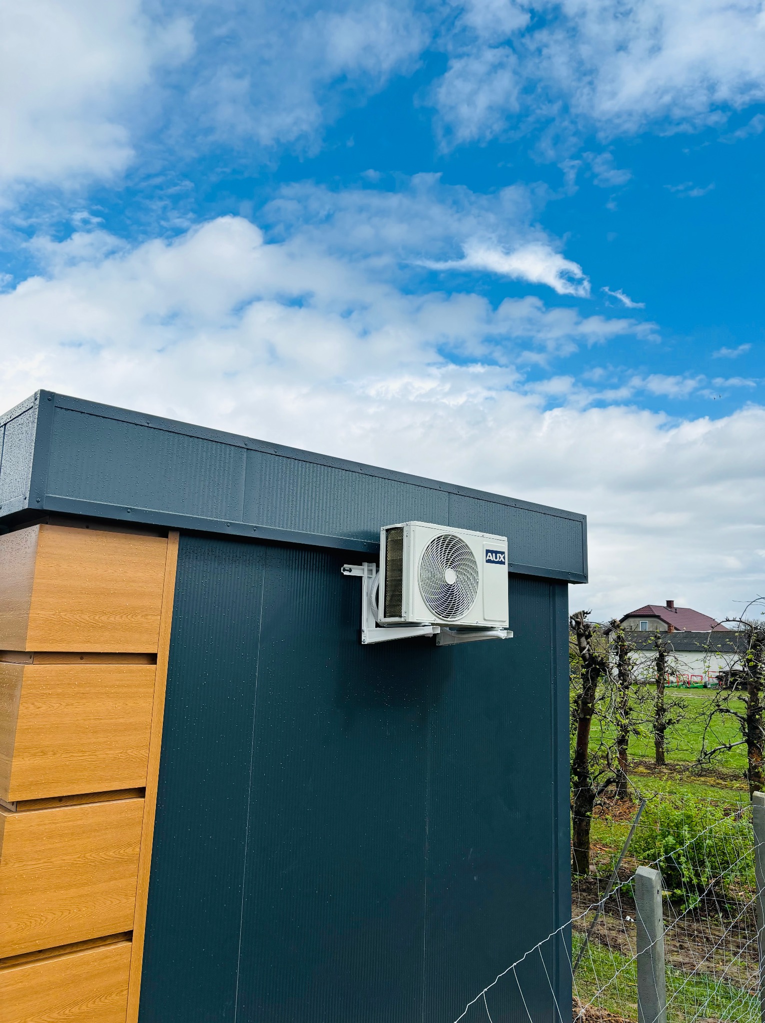 Klimatyzator AUX zamontowany na elewacji budynku z elementami drewnopodobnymi. W tle błękitne niebo z chmurami i zieleń ogrodu.