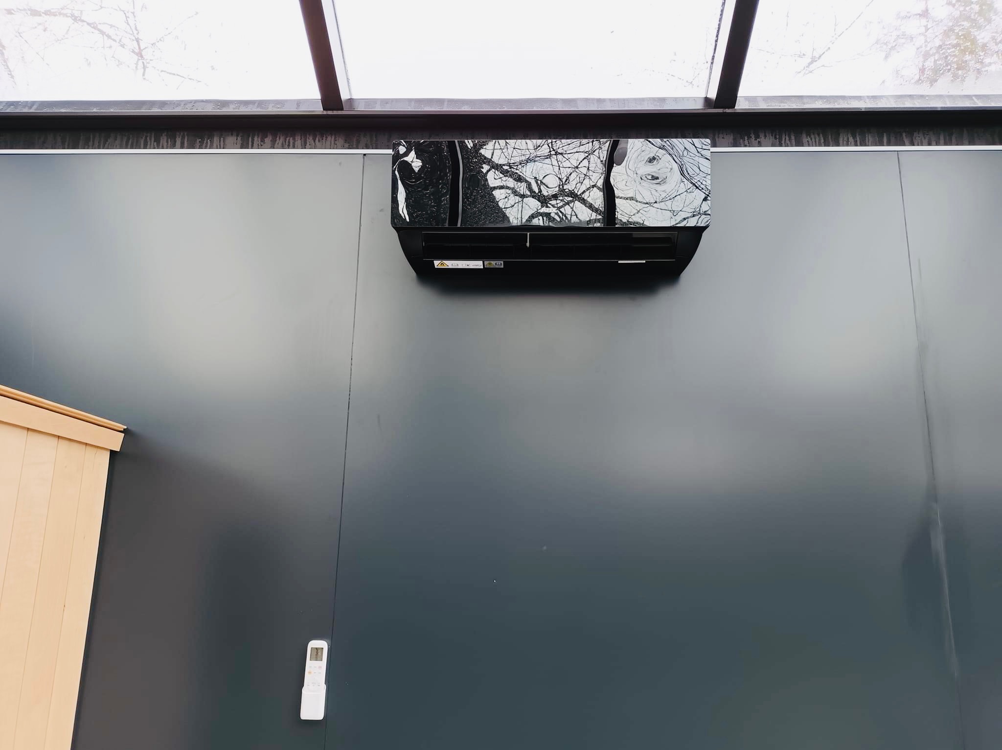 Zamontowana, czarna klimatyzacja na szarej ścianie pod przeszklonym dachem, z widocznym pilotem. W lustrzanej powierzchni odbija się drzewo.