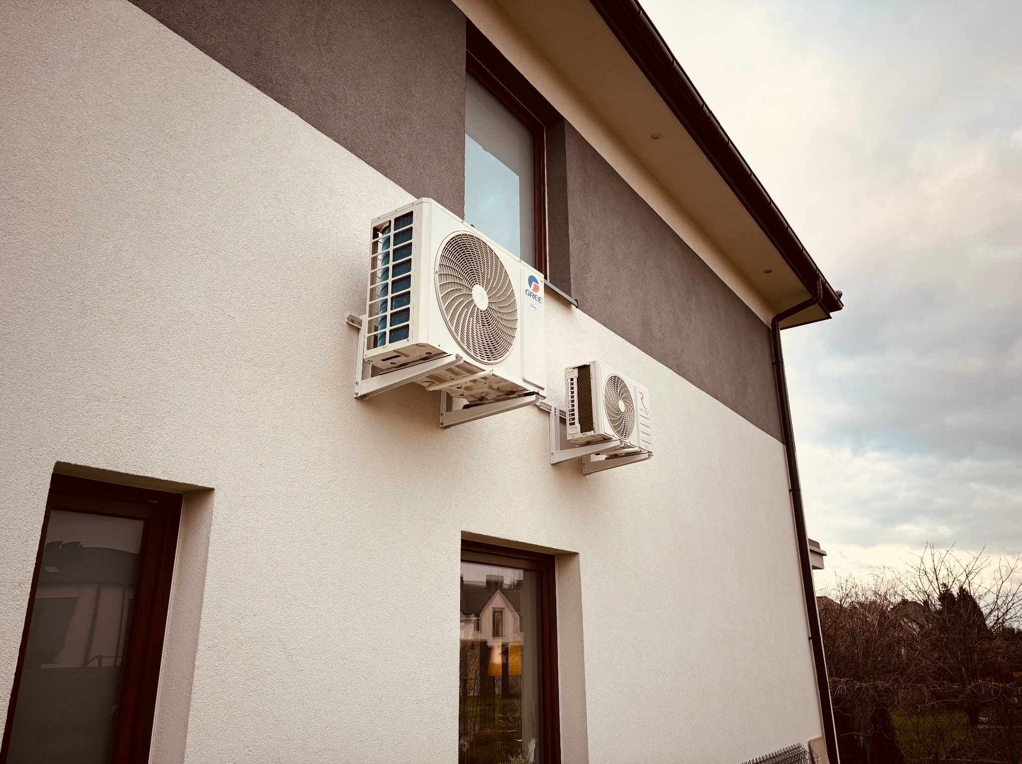 Dwa białe klimatyzatory Gree zamontowane na elewacji nowoczesnego domu z oknami. Widok z dołu na dwukolorową fasadę i pochmurne niebo.
