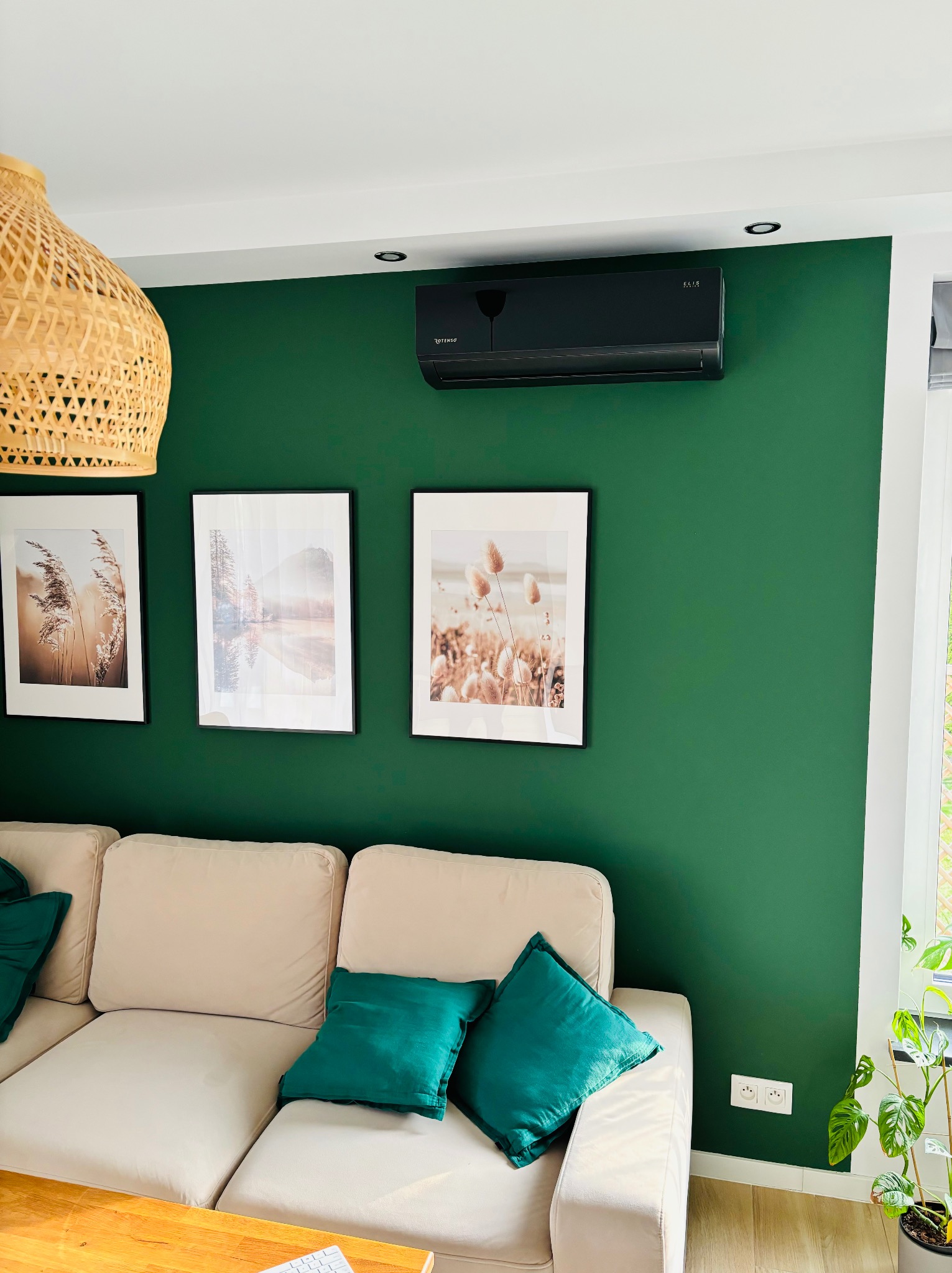 Nowoczesny, czarny klimatyzator na zielonej ścianie w salonie. Nad kanapą wiszą trzy obrazy w czarnych ramkach, a obok okna stoi zielona roślina doniczkowa.
