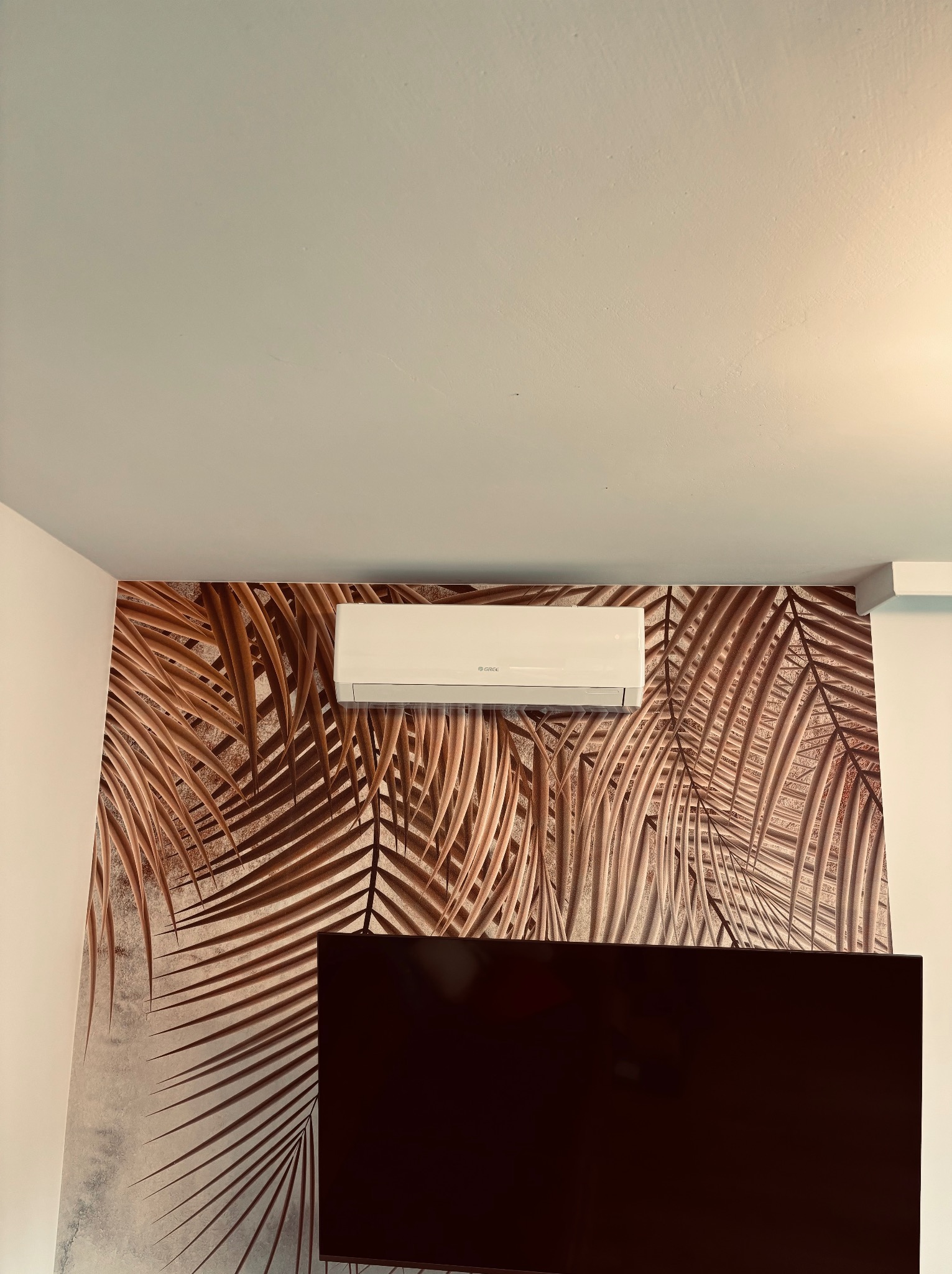 Biały klimatyzator Gree zamontowany na ścianie z tapetą w brązowe liście palmowe, widok z dołu, fragment telewizora w dolnej części kadru.