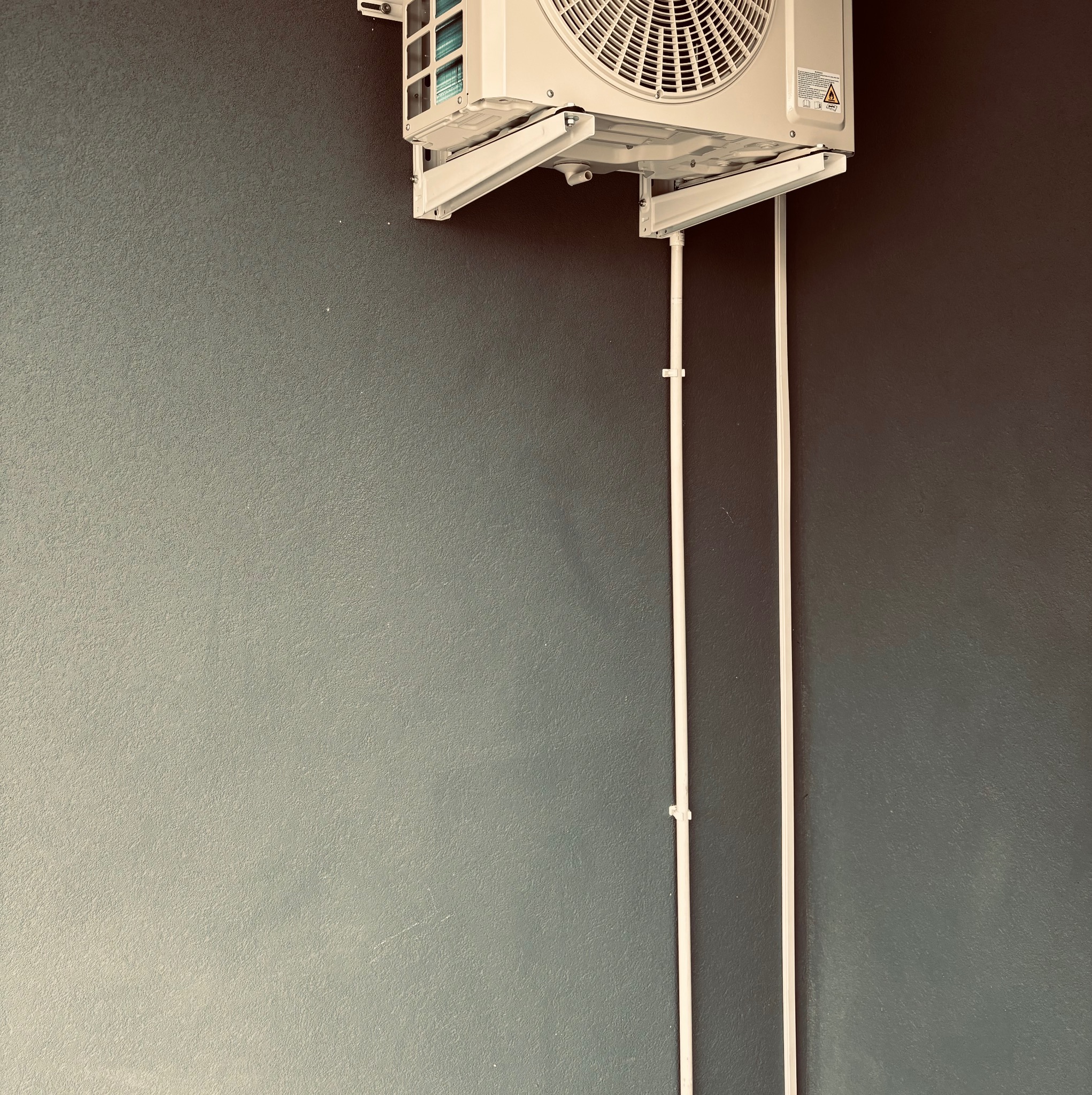 Biała jednostka klimatyzacji zamontowana na szarej ścianie. Dwie białe rurki prowadzą w dół od urządzenia. Widoczne elementy montażowe i ostrzeżenie na obudowie.