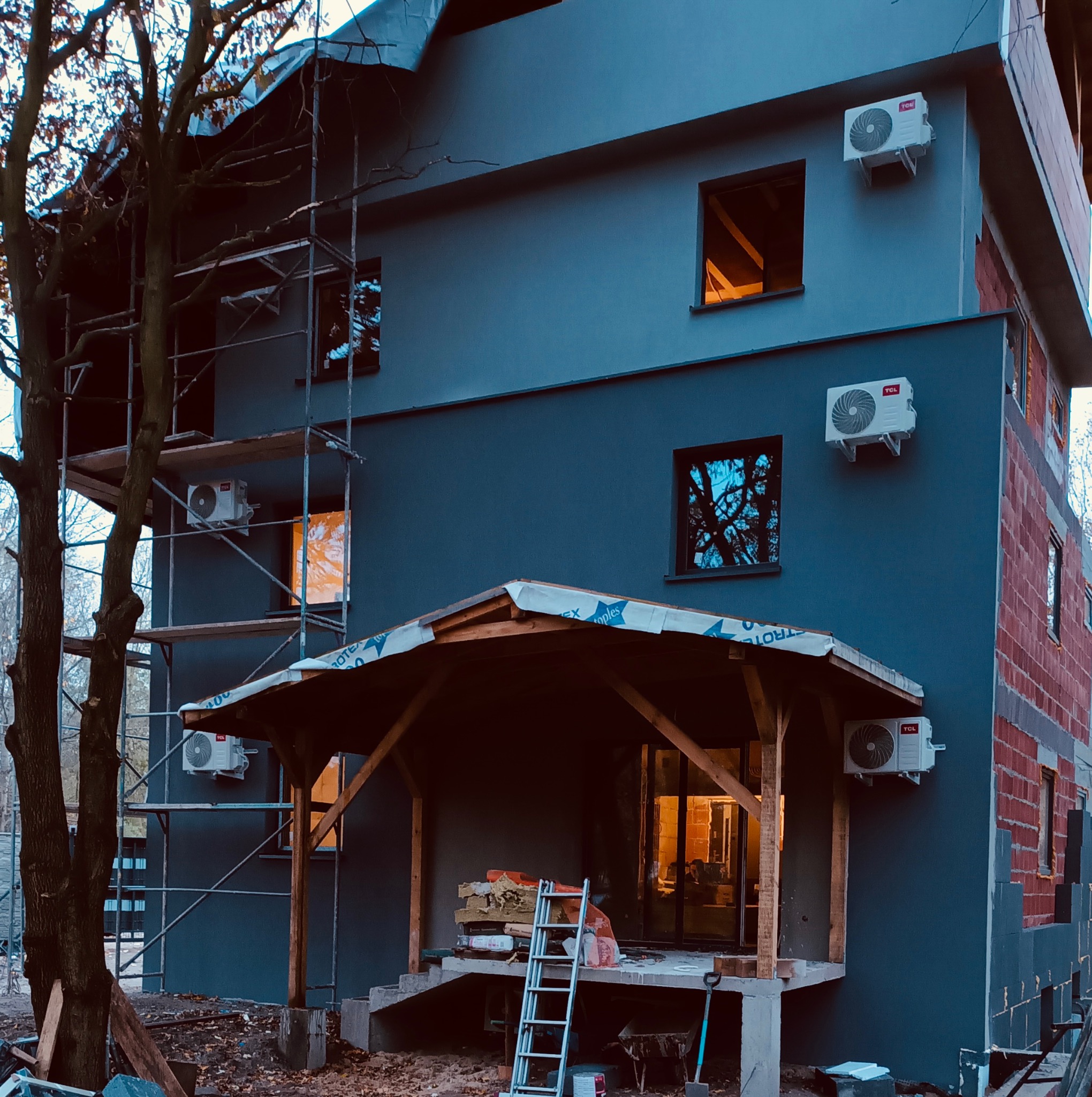 Elewacja domu w trakcie montażu klimatyzacji TCL. Widoczne rusztowania, okna z odbiciami i fragmenty niezakończonej elewacji z cegły. Częściowo oświetlone wnętrze.