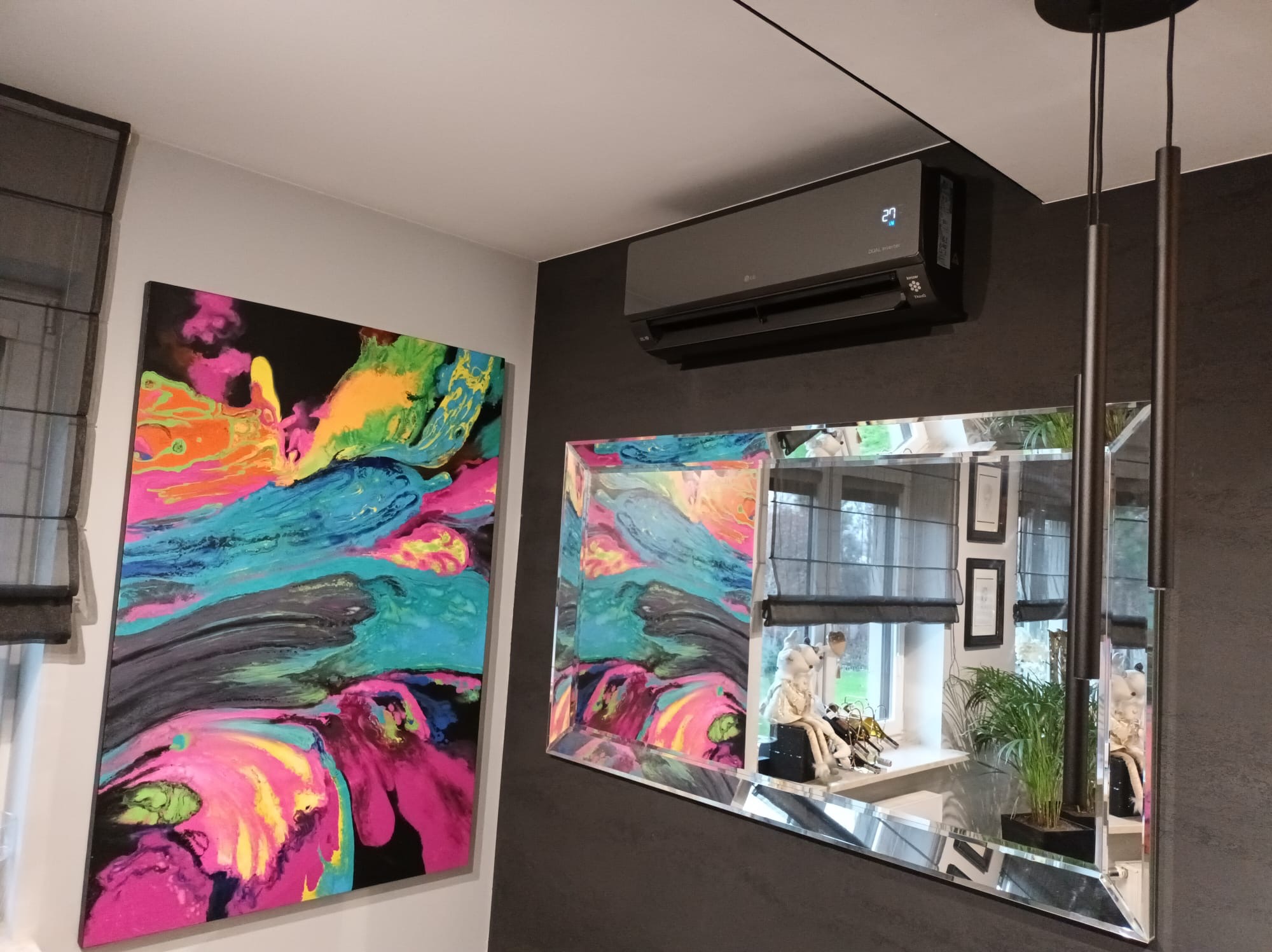 Nowoczesny klimatyzator LG w ciemnej obudowie, zamontowany w pokoju z obrazem i lustrem, oświetlony lampą wiszącą. Widoczny fragment okna z roletami.