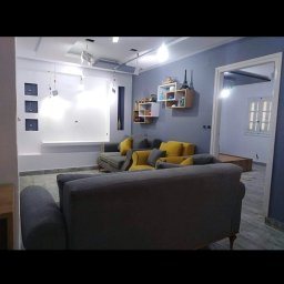 OSMANE BELAMRI - Szare ściany w salonie z szarymi sofami z żółtymi poduszkami, białe półki z dekoracjami i białe elementy dekoracyjne na ścianie.