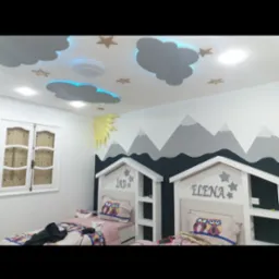 Pokój dziecięcy z dekoracyjnym malowaniem ścian przedstawiającym góry, oraz sufit z elementami chmur i gwiazd, podświetlony na niebiesko, dodatkowo dwa łóżka z zagłówkami w kształcie domków...