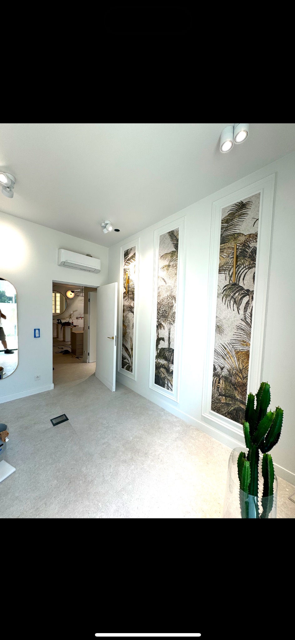 Wnętrze pokoju po remoncie z trzema pionowymi panelami dekoracyjnymi z motywem tropikalnych liści w białych ramach, jasna podłoga, widoczny kaktus w szklanym wazonie i otwarte drzwi do łazienki.