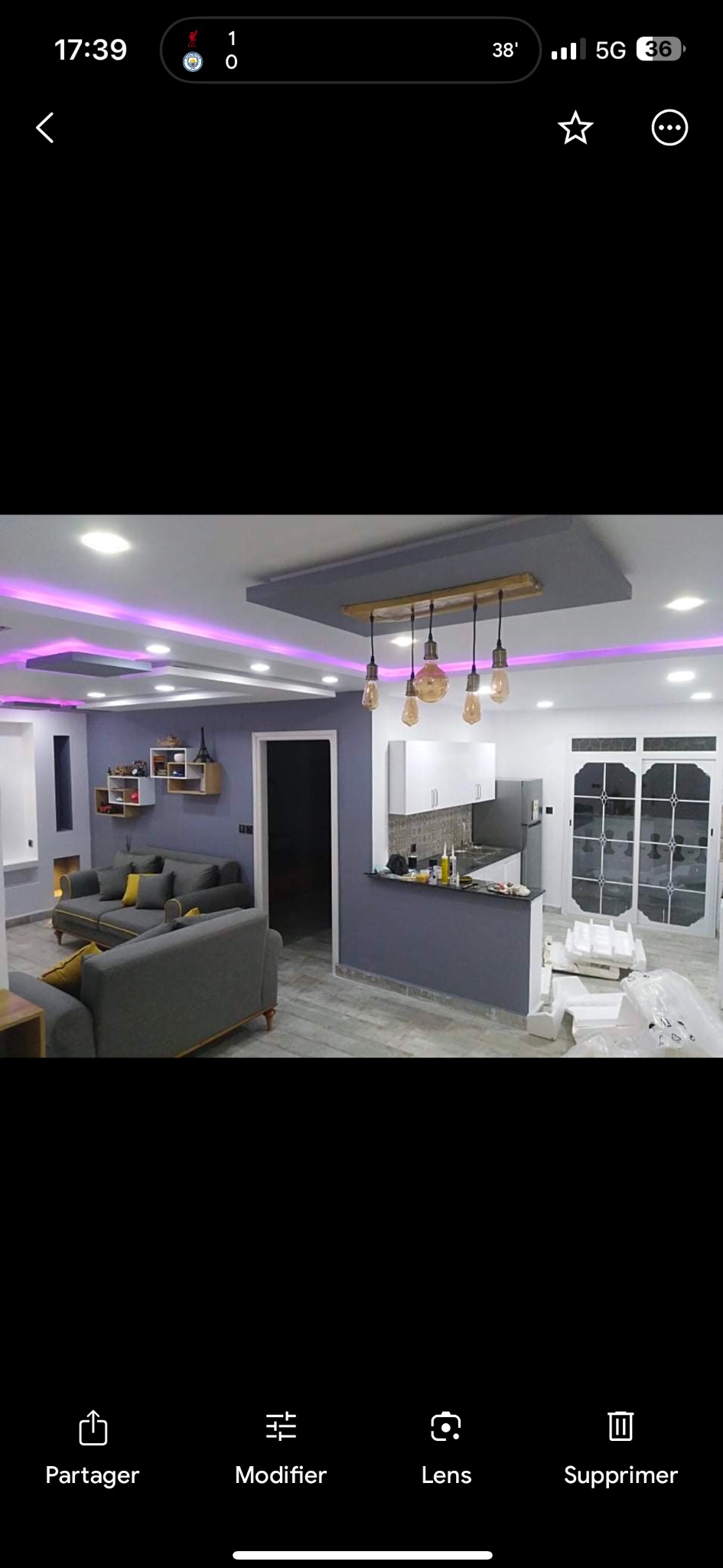 Nowoczesne wnętrze salonu z aneksem kuchennym, szare ściany, podwieszany sufit z oświetleniem LED, szara sofa z żółtymi poduszkami, widoczne elementy umeblowania kuchennego, częściowo rozpakowane...