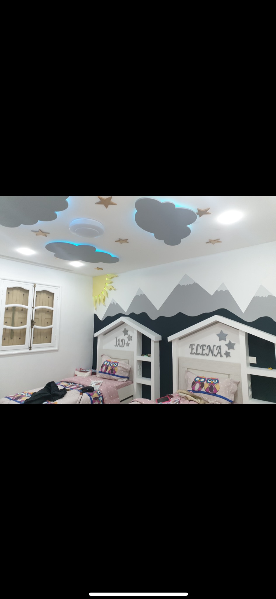 Pokój dziecięcy z dekoracyjnym malowaniem ścian przedstawiającym góry, oraz sufit z elementami chmur i gwiazd, podświetlony na niebiesko, dodatkowo dwa łóżka z zagłówkami w kształcie domków...