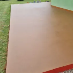 Beżowa, gładka powierzchnia wylewki na zewnątrz budynku, z widocznymi puszkami farb w tle, otoczona trawnikiem i zielenią.