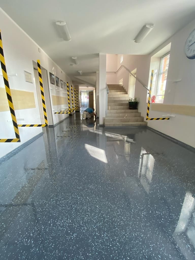 Szary korytarz w budynku publicznym z błyszczącą posadzką, zabezpieczony taśmą ostrzegawczą, pracownik w oddali poleruje podłogę.