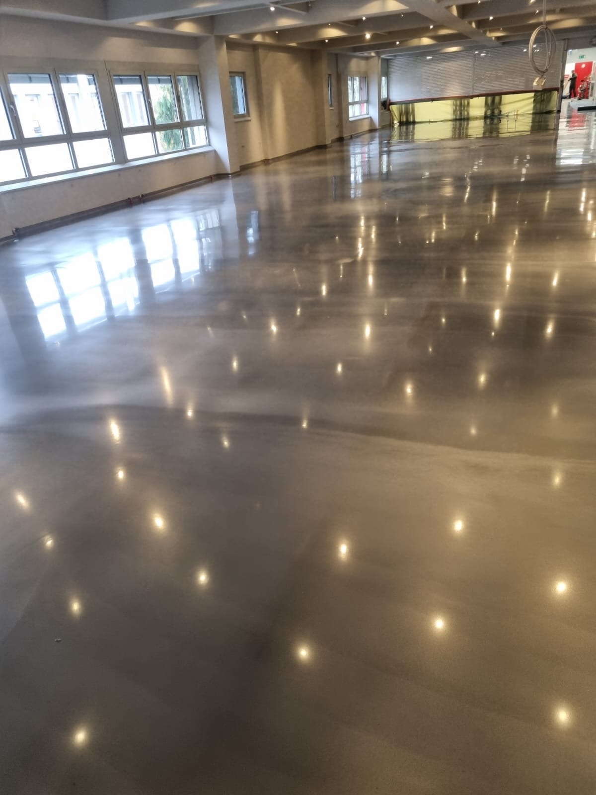 Wypolerowana, szara posadzka w przestronnym, jasnym pomieszczeniu. Refleksy świetlne od lamp i okien tworzą efekt lustrzanej powierzchni.