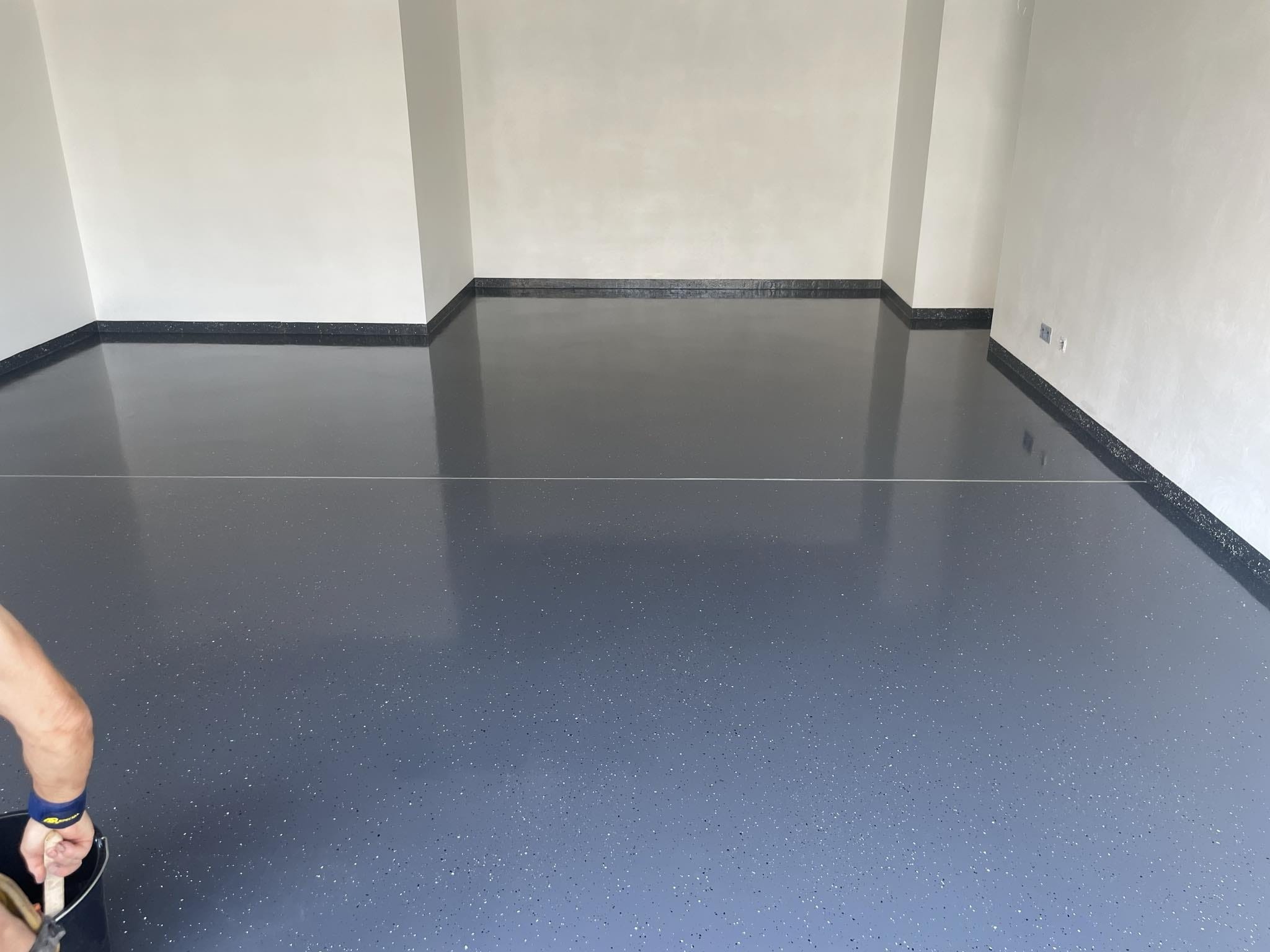 Błyszcząca, ciemnoszara posadzka z drobnymi, jasnymi wtrąceniami w pustym pomieszczeniu z białymi ścianami i ciemnym cokolikiem, widoczne narzędzie i ręka wykonawcy z boku.