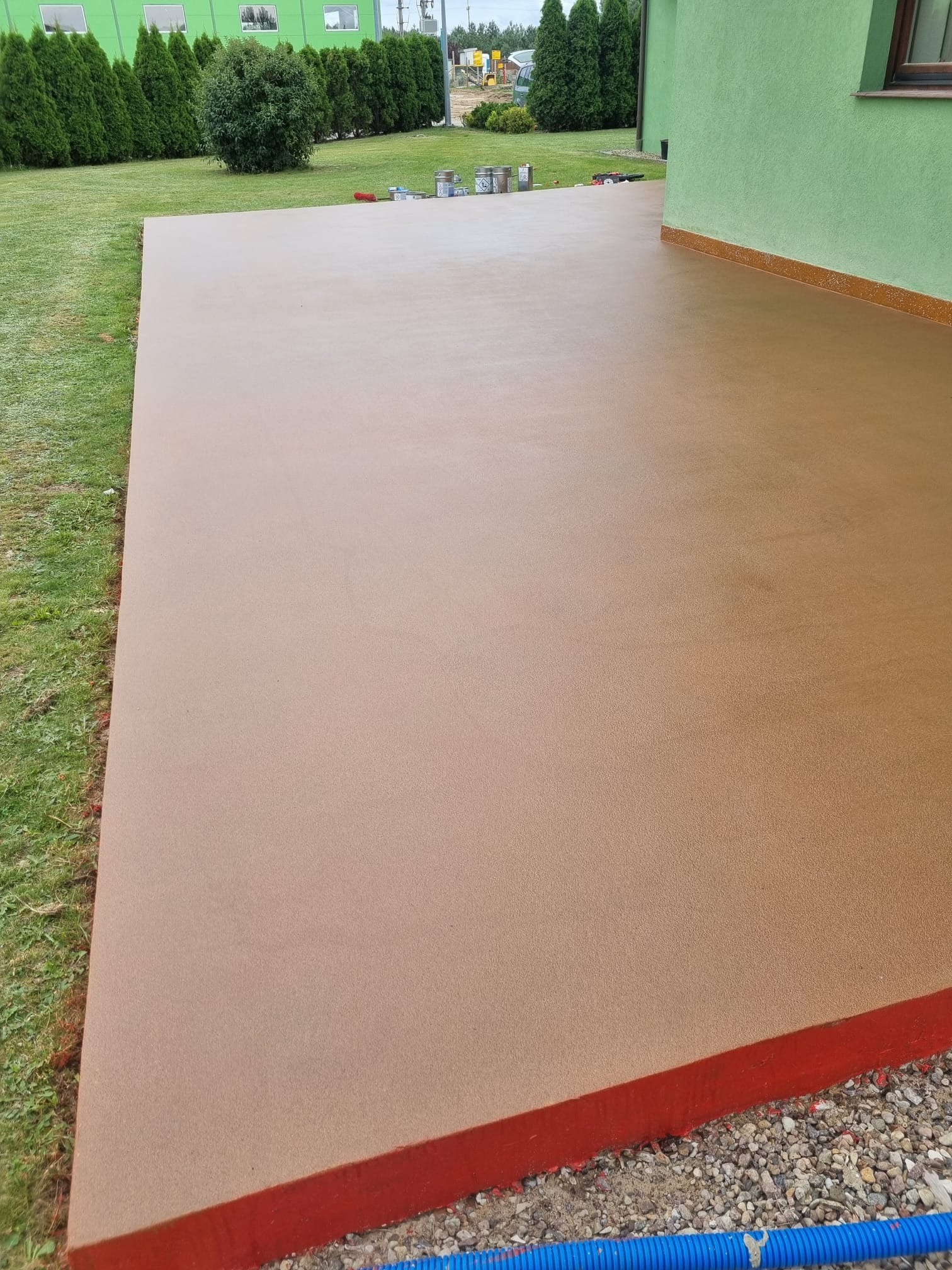 Beżowa, gładka powierzchnia wylewki na zewnątrz budynku, z widocznymi puszkami farb w tle, otoczona trawnikiem i zielenią.