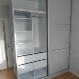 Kamil Wilgos - Biała szafa wnękowa z przesuwnymi drzwiami i otwartą częścią z półkami oraz szufladami, na drewnianej podłodze, widoczna biała lampa sufitowa.