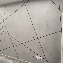 Kamil Wilgos - Szara ściana z dekoracyjnymi czarnymi liniami tworzącymi geometryczny wzór, widoczne gniazdka elektryczne i biały grzejnik.