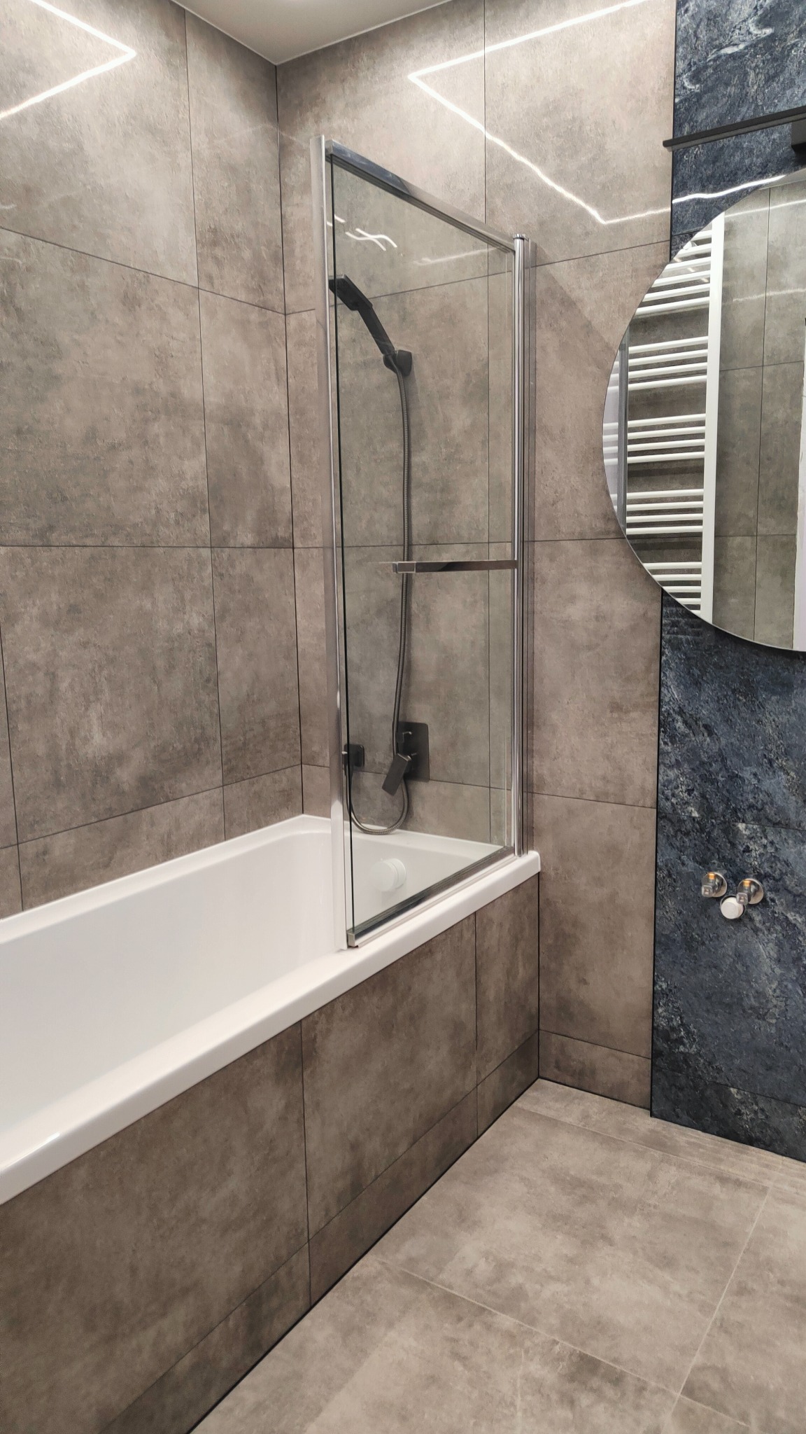 Nowoczesna łazienka z wanną i szklanym parawanem, wykończona płytkami w odcieniach szarości i granatu, z okrągłym lustrem i grzejnikiem drabinkowym.