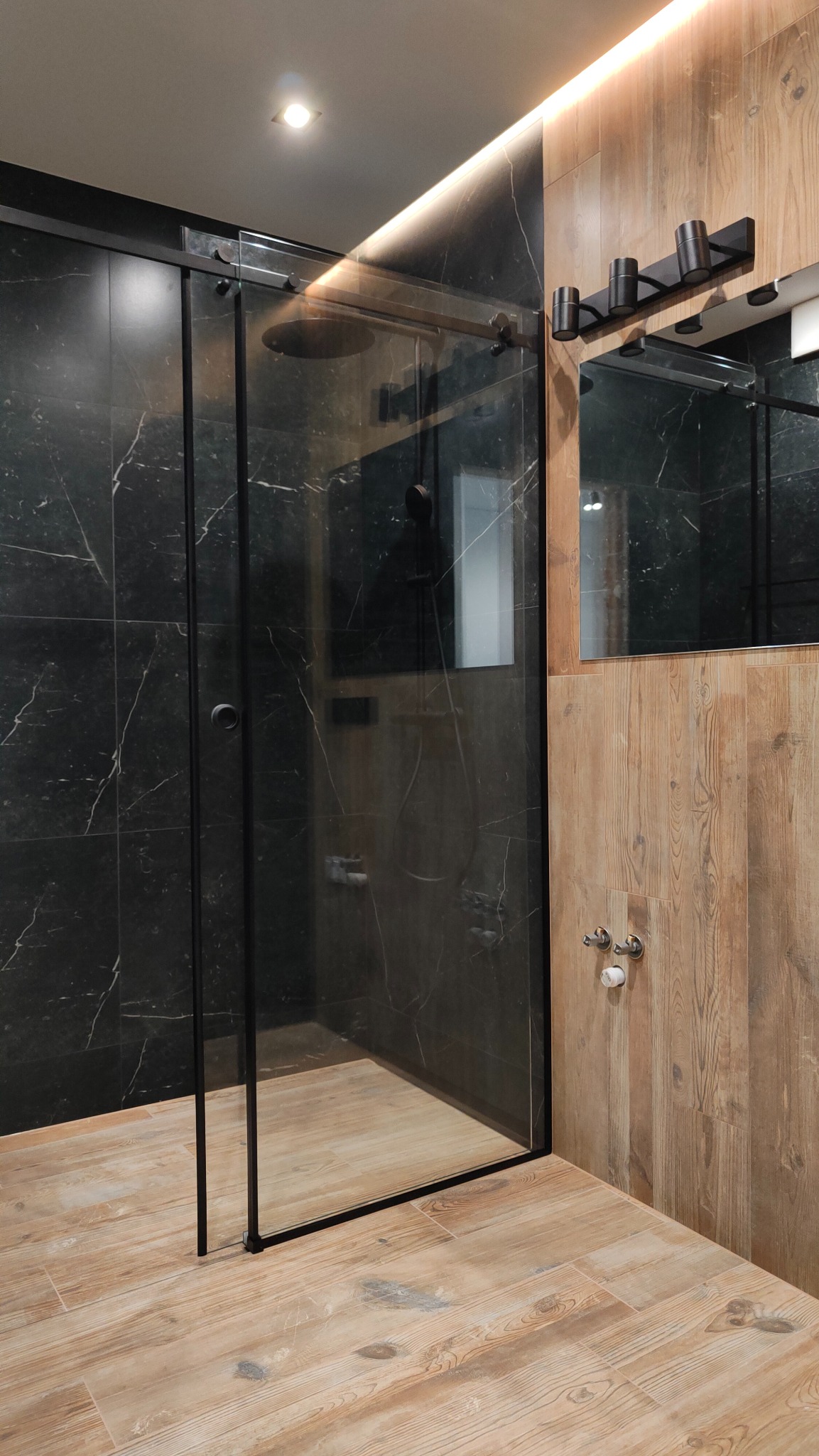 Nowoczesna kabina prysznicowa typu walk-in z czarną ramą, obudowana czarnymi płytkami imitującymi marmur i drewnopodobnymi panelami na ścianie, z widoczną armaturą i oświetleniem LED.