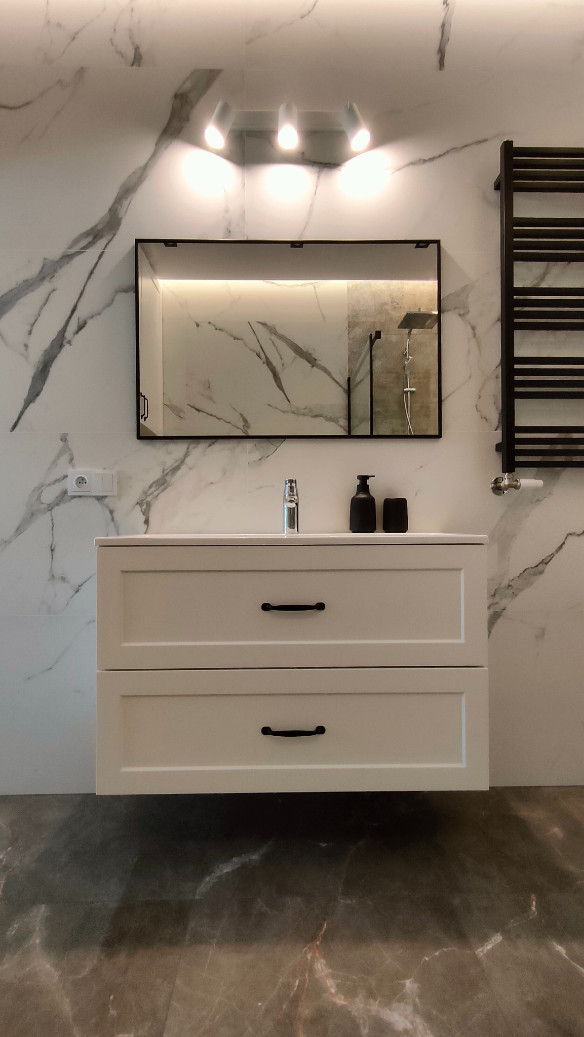 Nowoczesna łazienka z marmurowymi płytkami na ścianach i podłodze, biała szafka pod umywalkę z czarnymi uchwytami, lustro w czarnej ramie z oświetleniem LED i widocznym fragmentem kabiny...