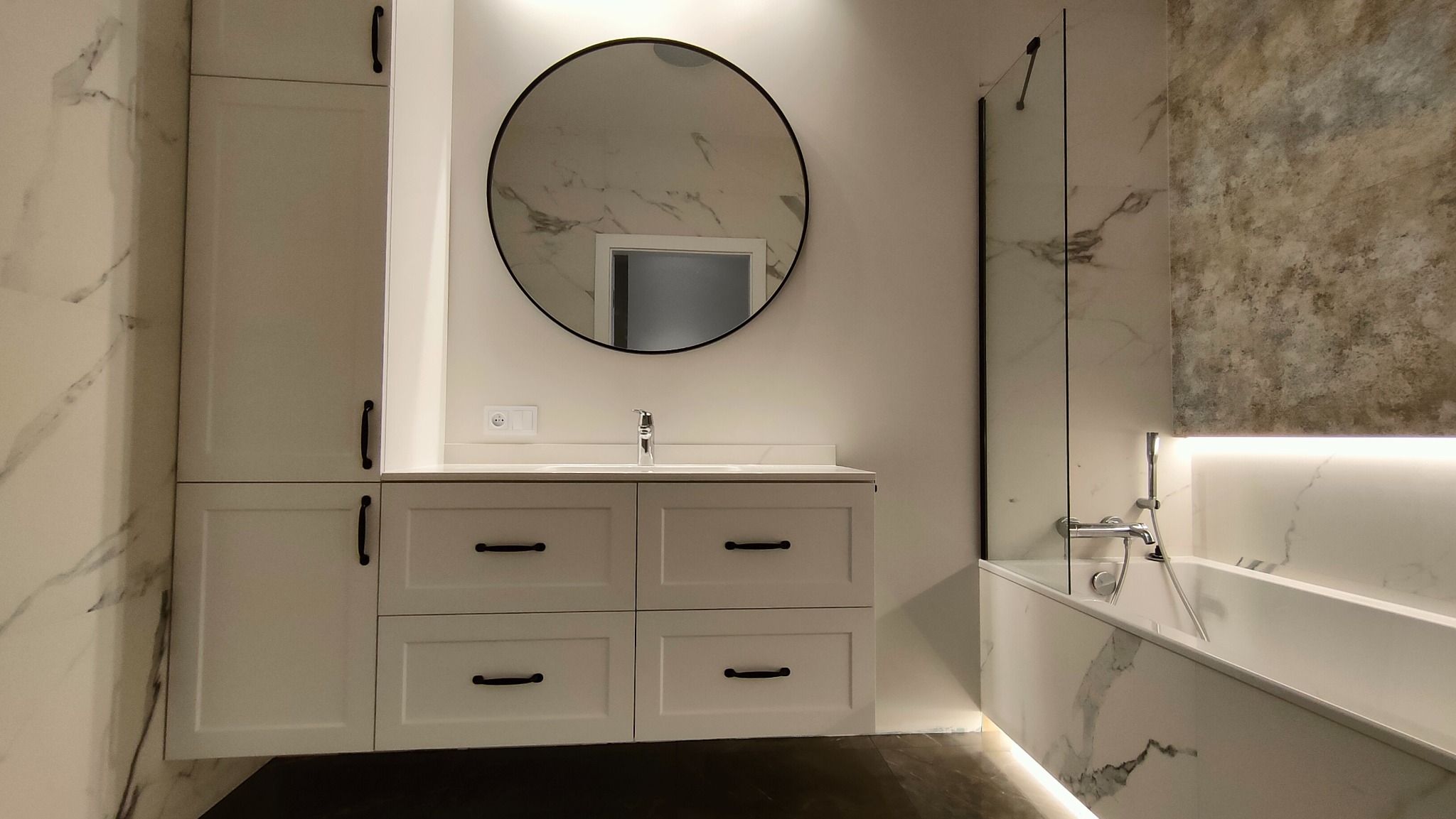 Nowoczesna łazienka z białą zabudową meblową, okrągłym lustrem z czarną ramą i wanną obudowaną marmurem, oświetlenie LED podkreśla marmurową fakturę ścian.