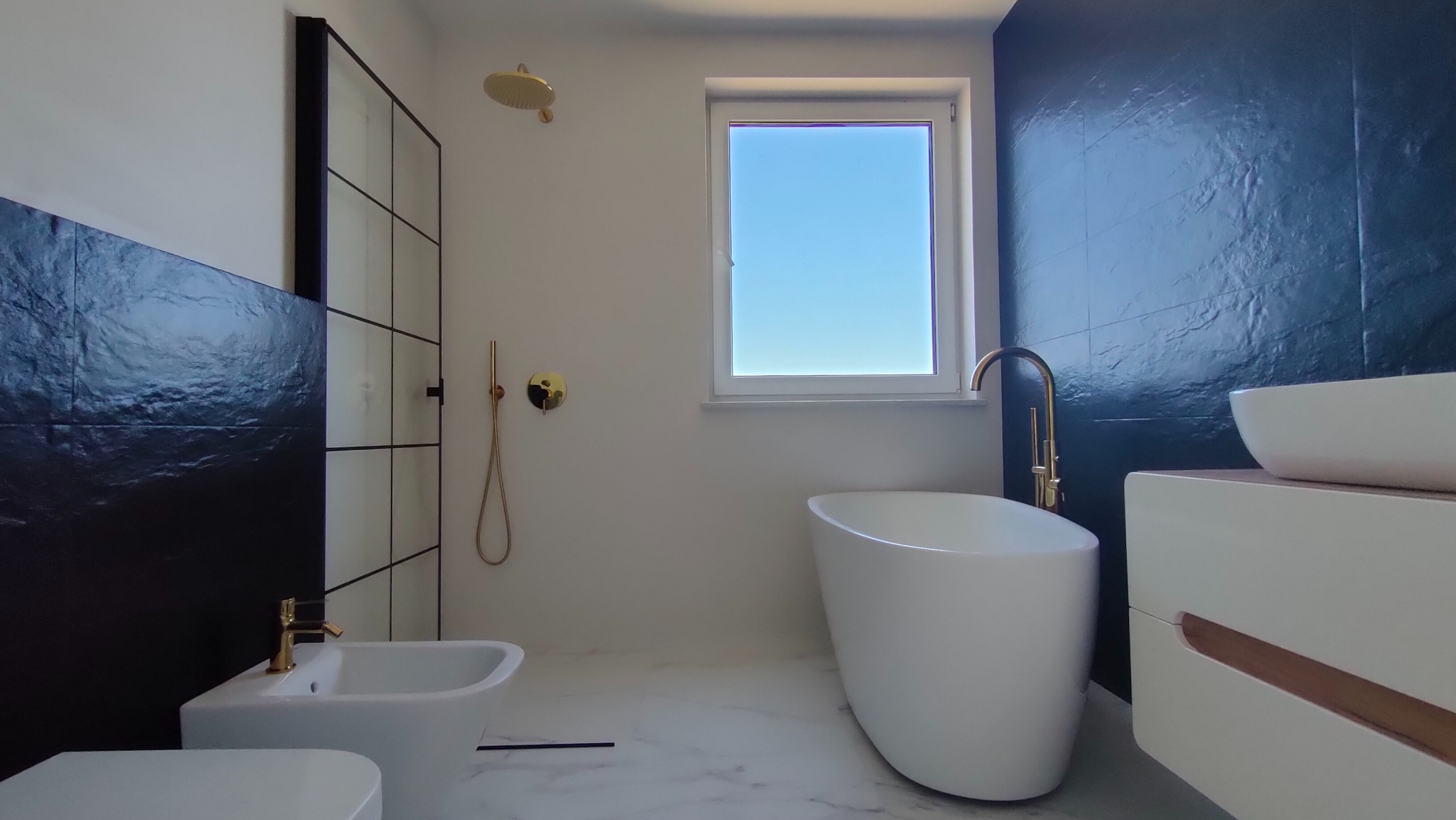 Nowoczesna łazienka z czarną ścianą z efektem kamienia, białą wolnostojącą wanną, bidetem i złotą armaturą, oświetlona naturalnym światłem z okna.