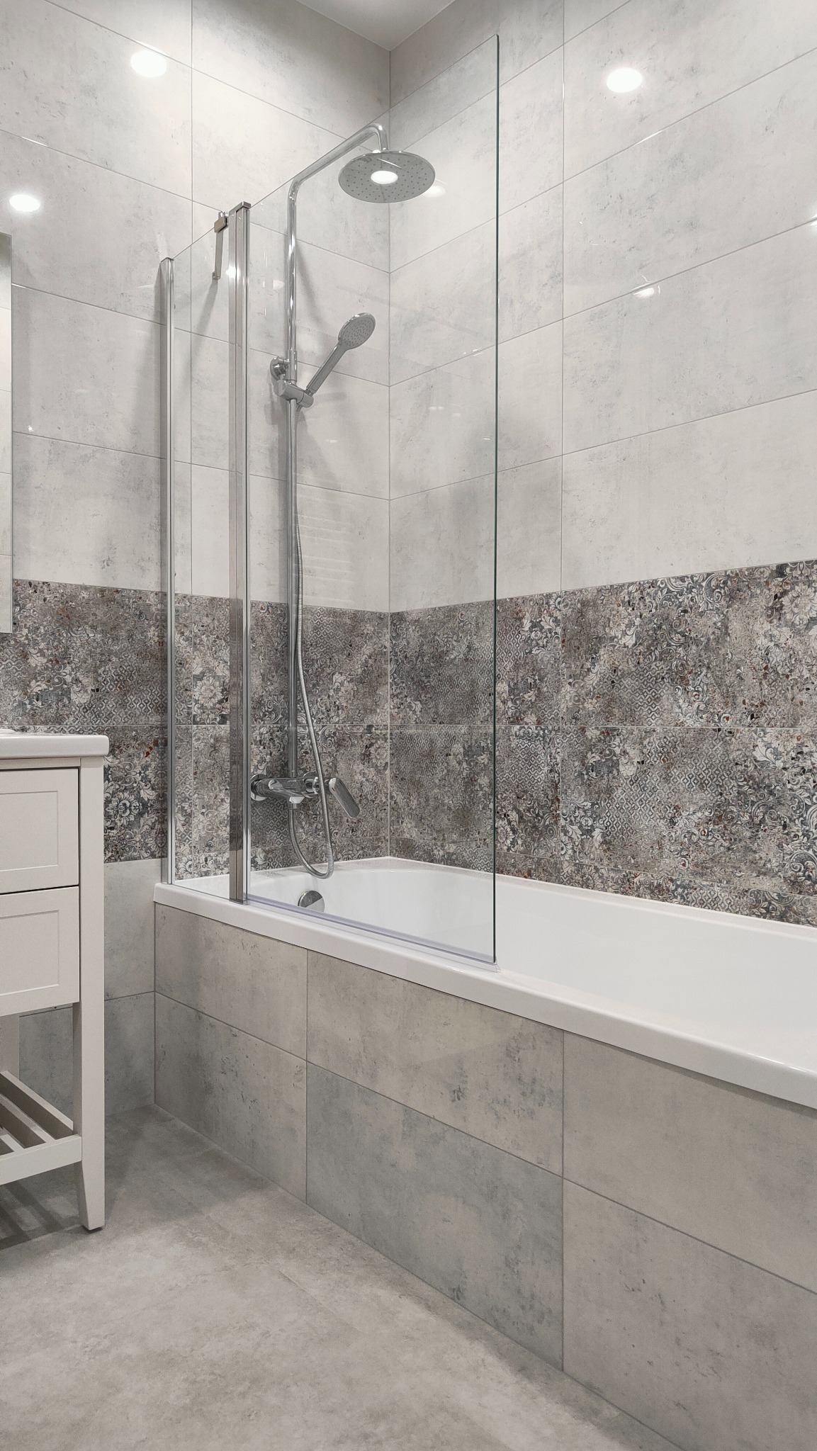 Nowoczesna łazienka z wanną i szklanym parawanem, wykończona płytkami w odcieniach szarości i bieli z dekoracyjnym pasem płytek wzorzystych, widoczny biały słupek umywalki.