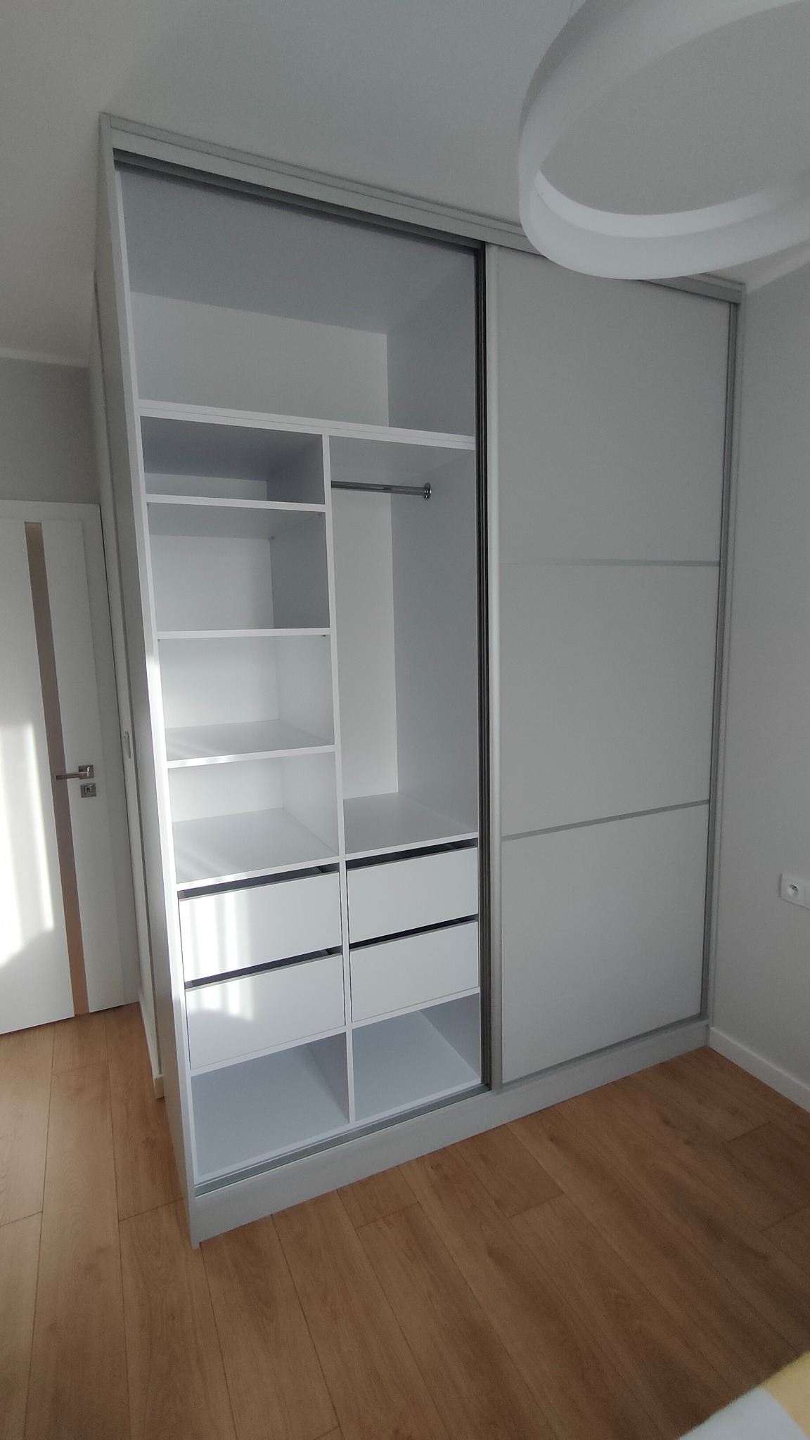 Biała szafa wnękowa z przesuwnymi drzwiami i otwartą częścią z półkami oraz szufladami, na drewnianej podłodze, widoczna biała lampa sufitowa.
