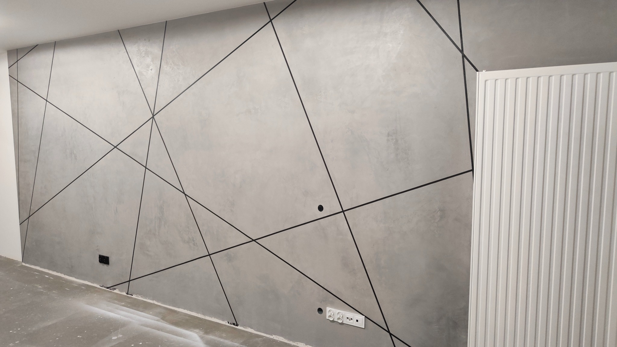 Szara ściana z dekoracyjnymi czarnymi liniami tworzącymi geometryczny wzór, widoczne gniazdka elektryczne i biały grzejnik.