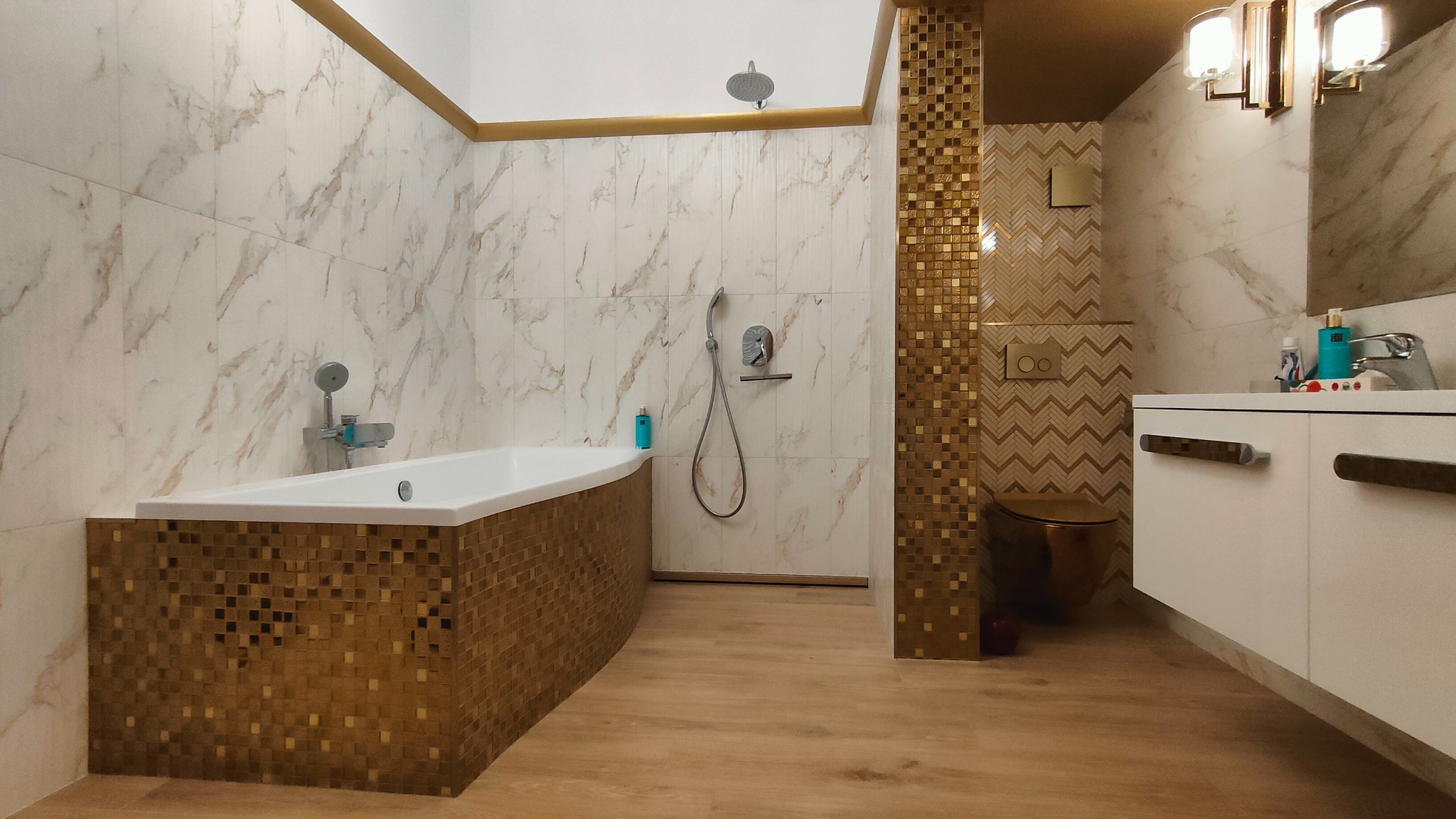Nowoczesna łazienka z wanną obudowaną mozaiką w odcieniach złota, marmurowe płytki na ścianach z subtelnymi złotymi akcentami, wisząca szafka z umywalką oraz toaleta w kolorze złotym.