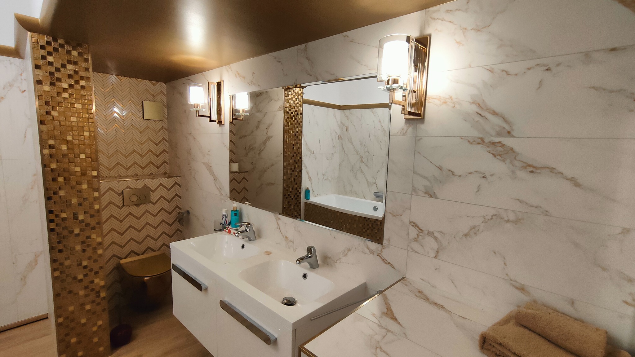Elegancka łazienka z marmurowymi płytkami, złotymi akcentami mozaiki i armaturą, widoczne dwie umywalki, lustro z oświetleniem i fragment toalety w kolorze złota.