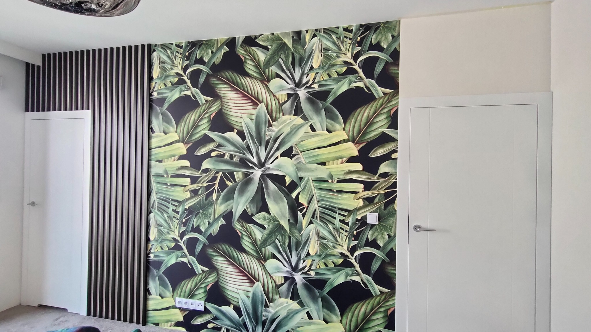 Nowoczesne wykończenie wnętrza z tapetą w tropikalne liście, dekoracyjnymi lamelami ściennymi i dwiema białymi drzwiami.
