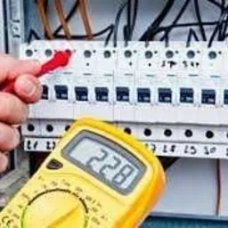 Miernik napięcia w dłoni, czerwony przewód dotyka bezpiecznika w skrzynce elektrycznej. Widoczne kable i oznaczenia na bezpiecznikach.