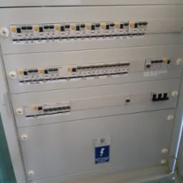 Rozdzielnia elektryczna z bezpiecznikami EATON, widoczne etykiety opisujące obwody. Szara obudowa rozdzielnicy na tle zielonej ściany. Widoczny wyłącznik główny.