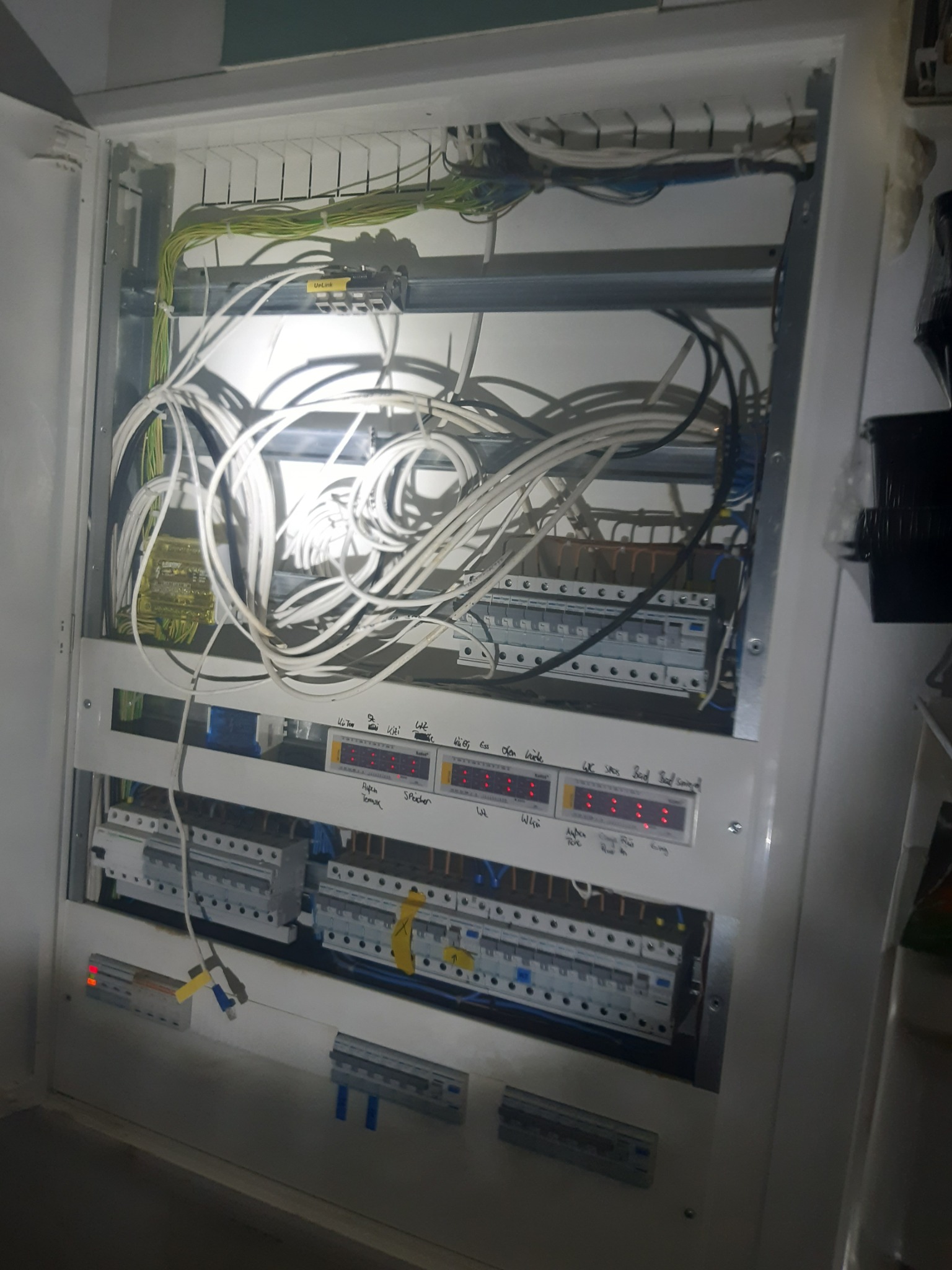 Wewnętrzny widok skrzynki rozdzielczej z licznymi przewodami, bezpiecznikami i wyświetlaczami. Widoczne opisy odręczne na panelu.