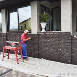 Valeriy - Pracownik w czerwonej koszulce i niebieskich spodniach, w słomkowym kapeluszu, układa ciemną cegłę na elewacji budynku, obok stoi czerwona piła do cięcia cegieł, na ziemi rozłożona folia ochronna.