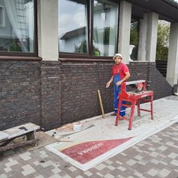 Valeriy - Pracownik w czerwonej koszulce i słomkowym kapeluszu tnie płytki elewacyjne na czerwonej przecinarce elektrycznej przed budynkiem z oknami, wykończonymi ciemnobrązową cegłą klinkierową.