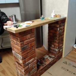 Valeriy - Konstrukcja z cegieł imitująca kominek w trakcie budowy, z drewnianym blatem, narzędziami i akcesoriami na blacie oraz wewnątrz wnęki, na podłodze kartonowe opakowanie.